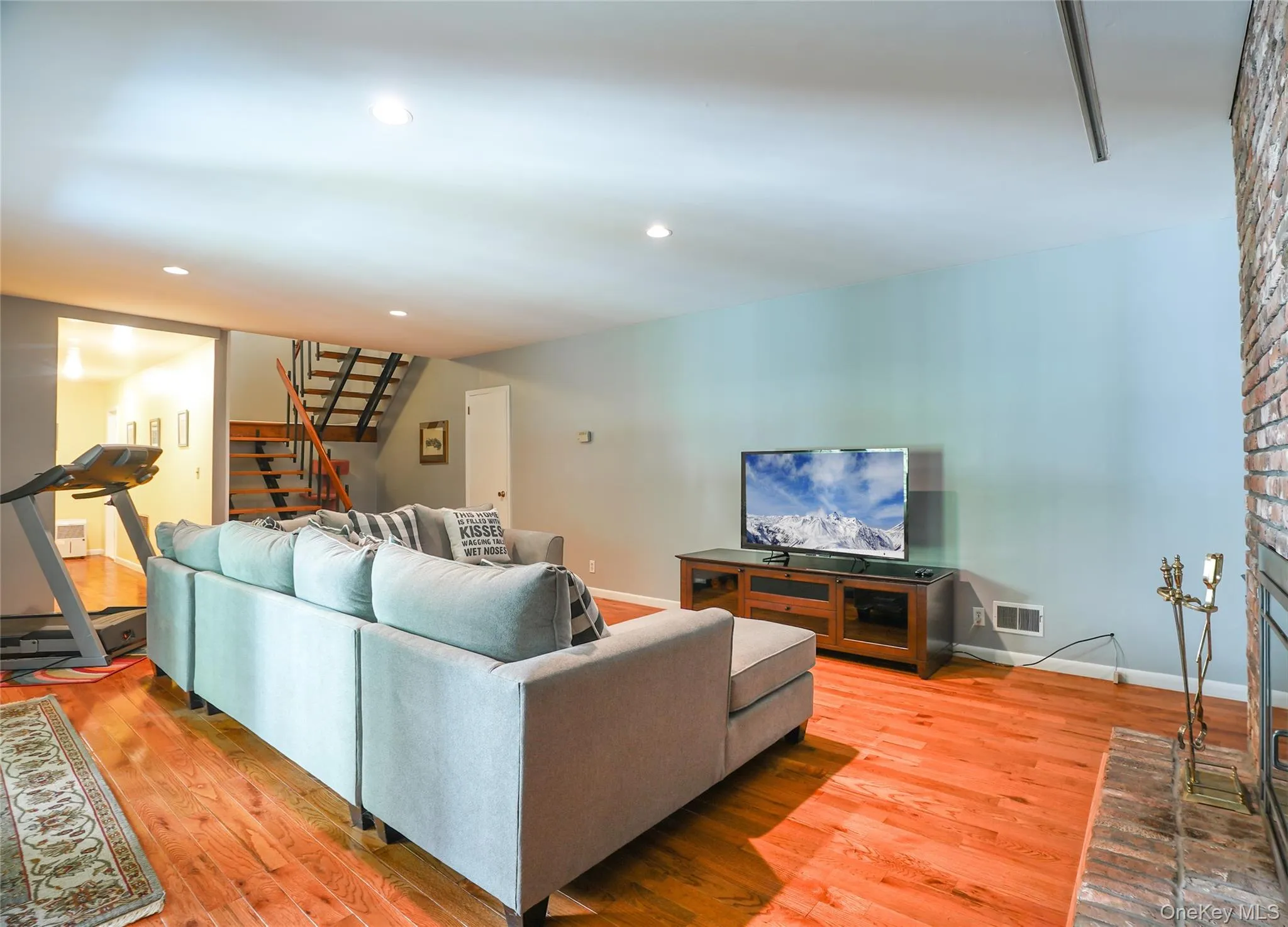 Spacious living area featuring wood-finish flooring, a full brick fireplace, recessed lighting, and an open-tread staircase Spacious living area featuring wood-finish flooring, a full brick fireplace, recessed lighting, and an open-tread staircase
