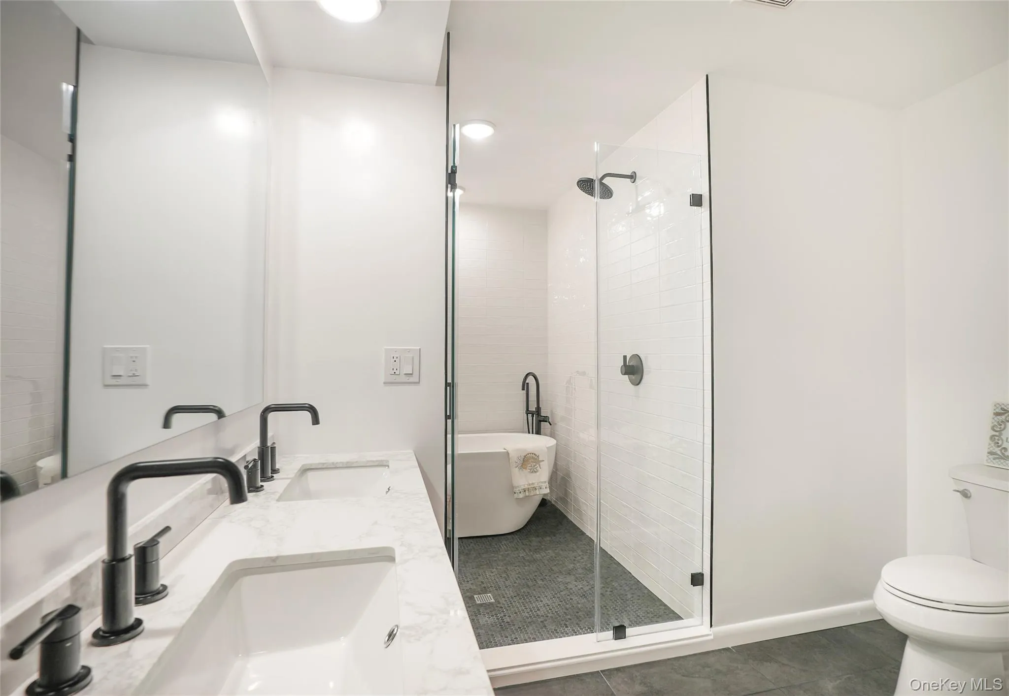 Spacious bathroom featuring a dual vanity with white countertop, matte black fixtures, a walk-in shower with clear glass enclosure, a freestanding soaking tub, and a toilet Spacious bathroom featuring a dual vanity with white countertop, matte black fixtures, a walk-in shower with clear glass enclosure, a freestanding soaking tub, and a toilet