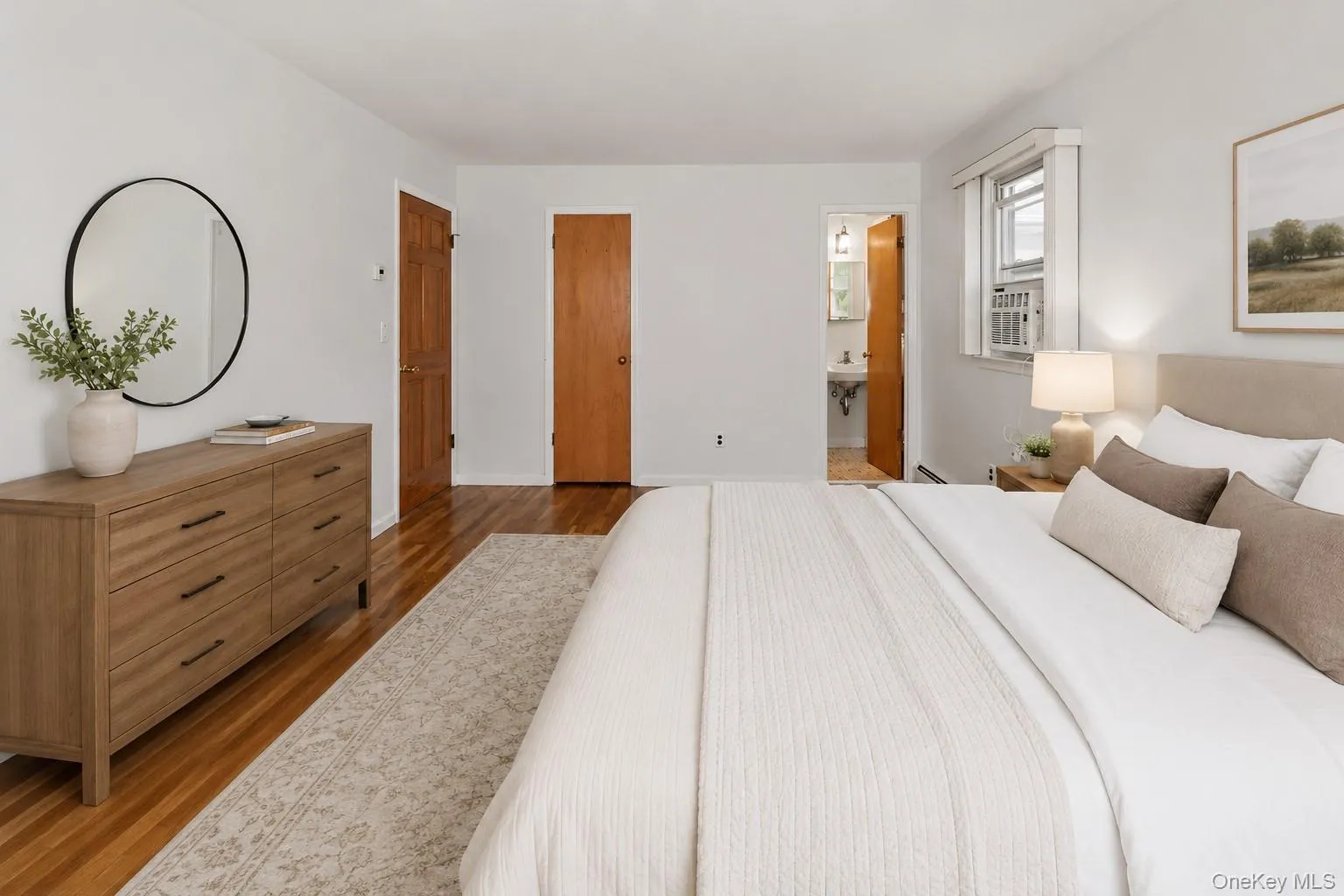 Virtually staged spacious bedroom featuring warm wood-finish flooring, light-colored walls, and a window with a built-in air conditioning unit Virtually staged spacious bedroom featuring warm wood-finish flooring, light-colored walls, and a window with a built-in air conditioning unit