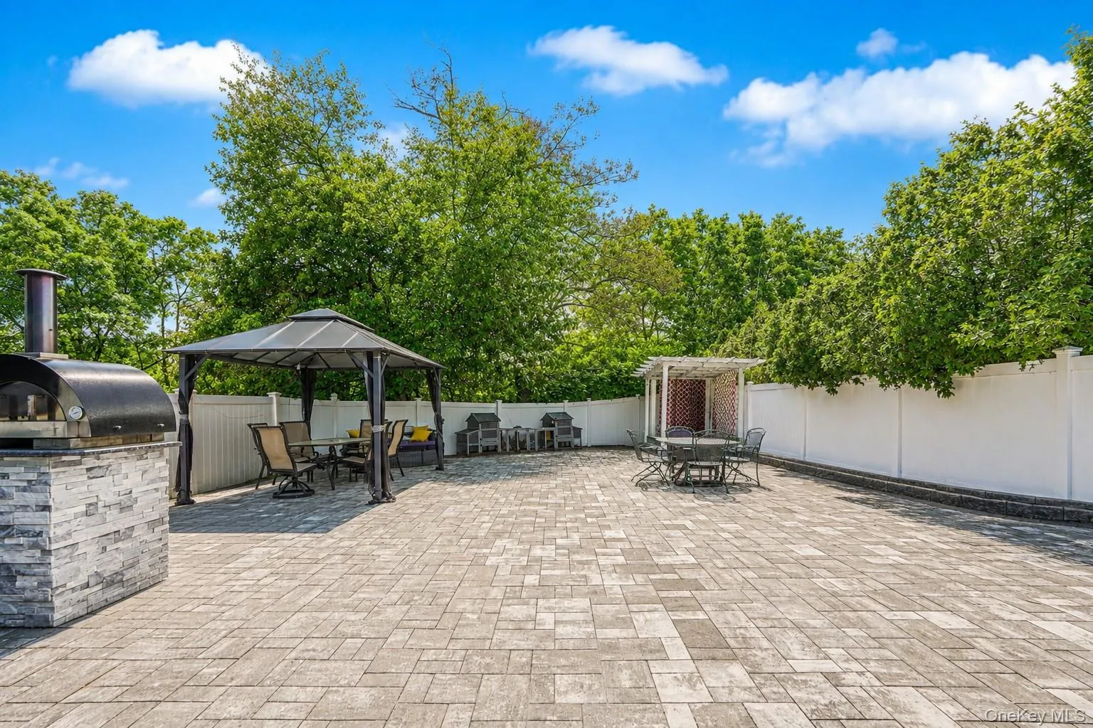 Expansive paver patio with a built-in outdoor pizza oven, a white privacy fence, and mature perimeter trees Expansive paver patio with a built-in outdoor pizza oven, a white privacy fence, and mature perimeter trees