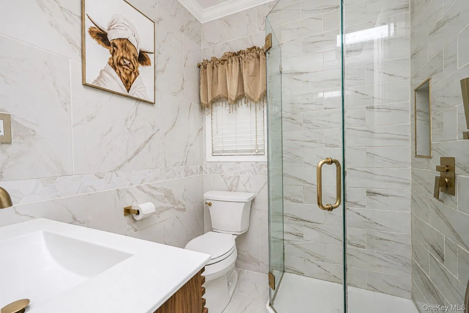 Bathroom featuring a glass-enclosed shower, white tile with subtle veining, gold-toned fixtures, a built-in shower niche, and a white vanity with an integrated sink Bathroom featuring a glass-enclosed shower, white tile with subtle veining, gold-toned fixtures, a built-in shower niche, and a white vanity with an integrated sink