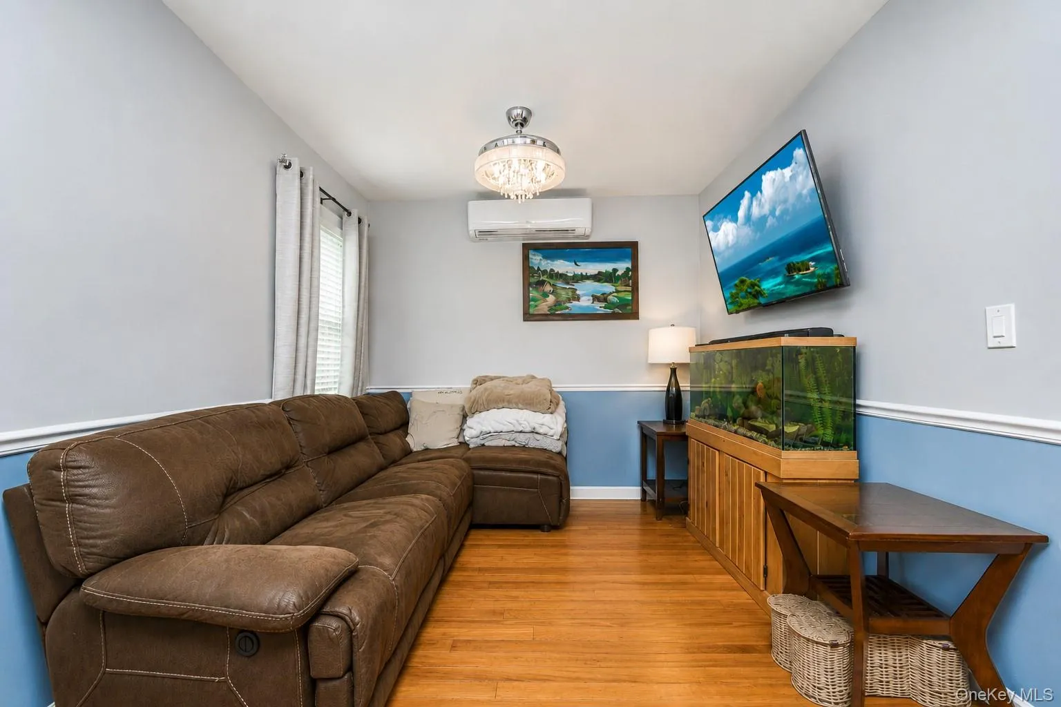 Living area featuring hardwood flooring, a two-tone wall design, a window with vertical blinds, a modern ceiling light fixture, and a wall-mounted mini-split system Living area featuring hardwood flooring, a two-tone wall design, a window with vertical blinds, a modern ceiling light fixture, and a wall-mounted mini-split system