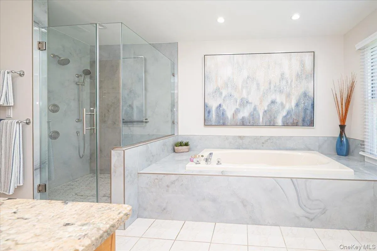 Spacious bathroom featuring a glass-enclosed shower with multiple showerheads, a built-in soaking tub, extensive light-toned stone-finish tiling, recessed lighting, and a window with blinds Spacious bathroom featuring a glass-enclosed shower with multiple showerheads, a built-in soaking tub, extensive light-toned stone-finish tiling, recessed lighting, and a window with blinds