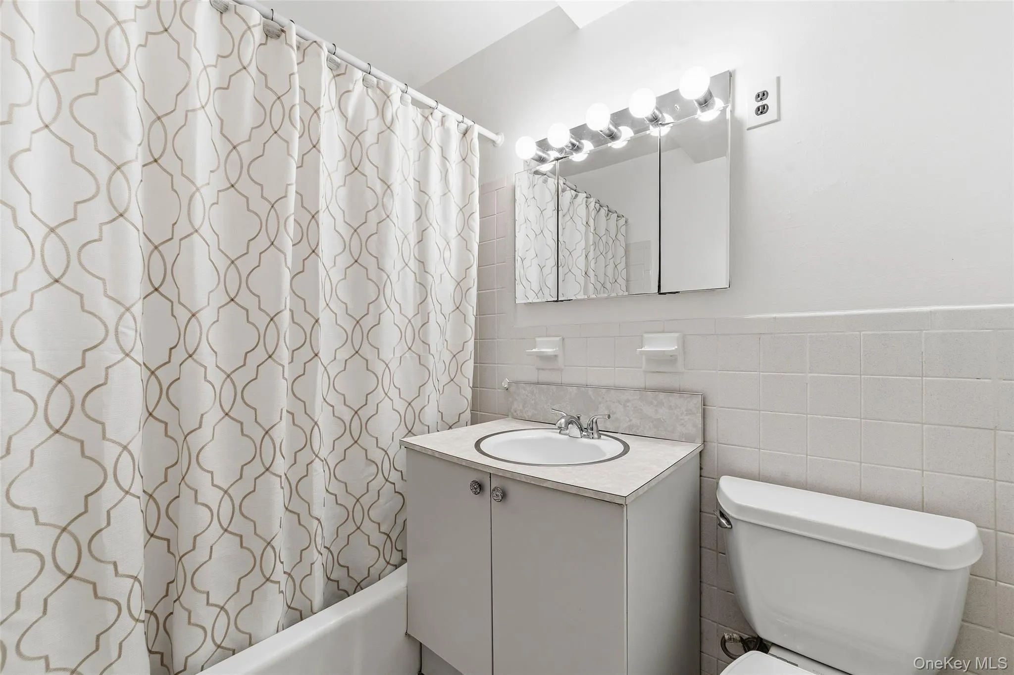 Bathroom featuring a white vanity with an integrated sink, a two-door medicine cabinet with mirrored fronts, a six-bulb vanity light fixture, a toilet, and white tiled walls Bathroom featuring a white vanity with an integrated sink, a two-door medicine cabinet with mirrored fronts, a six-bulb vanity light fixture, a toilet, and white tiled walls
