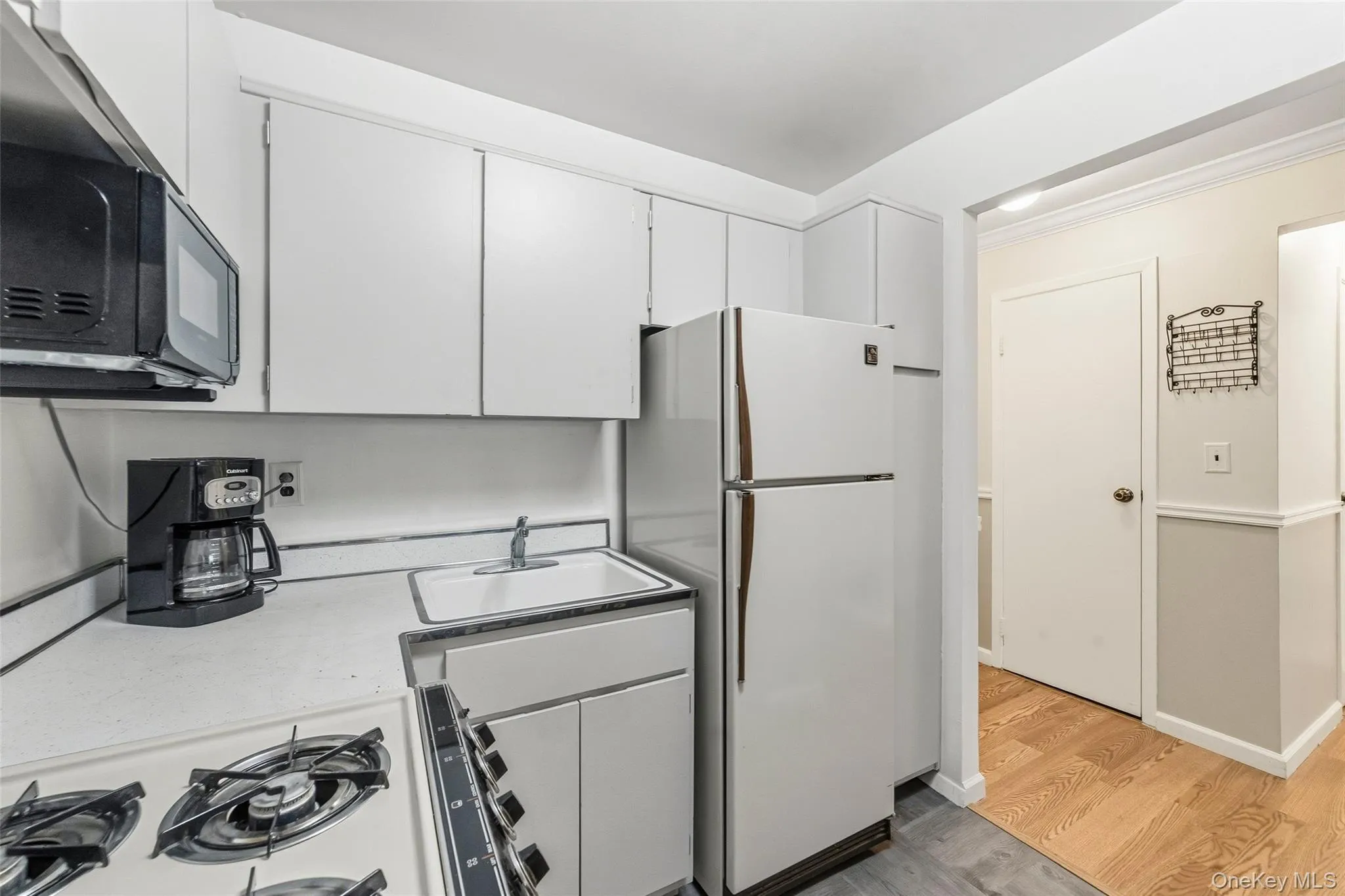 Kitchen featuring white cabinetry, a built-in white sink, a wall-mounted microwave, white refrigerator, and wood-finish flooring Kitchen featuring white cabinetry, a built-in white sink, a wall-mounted microwave, white refrigerator, and wood-finish flooring