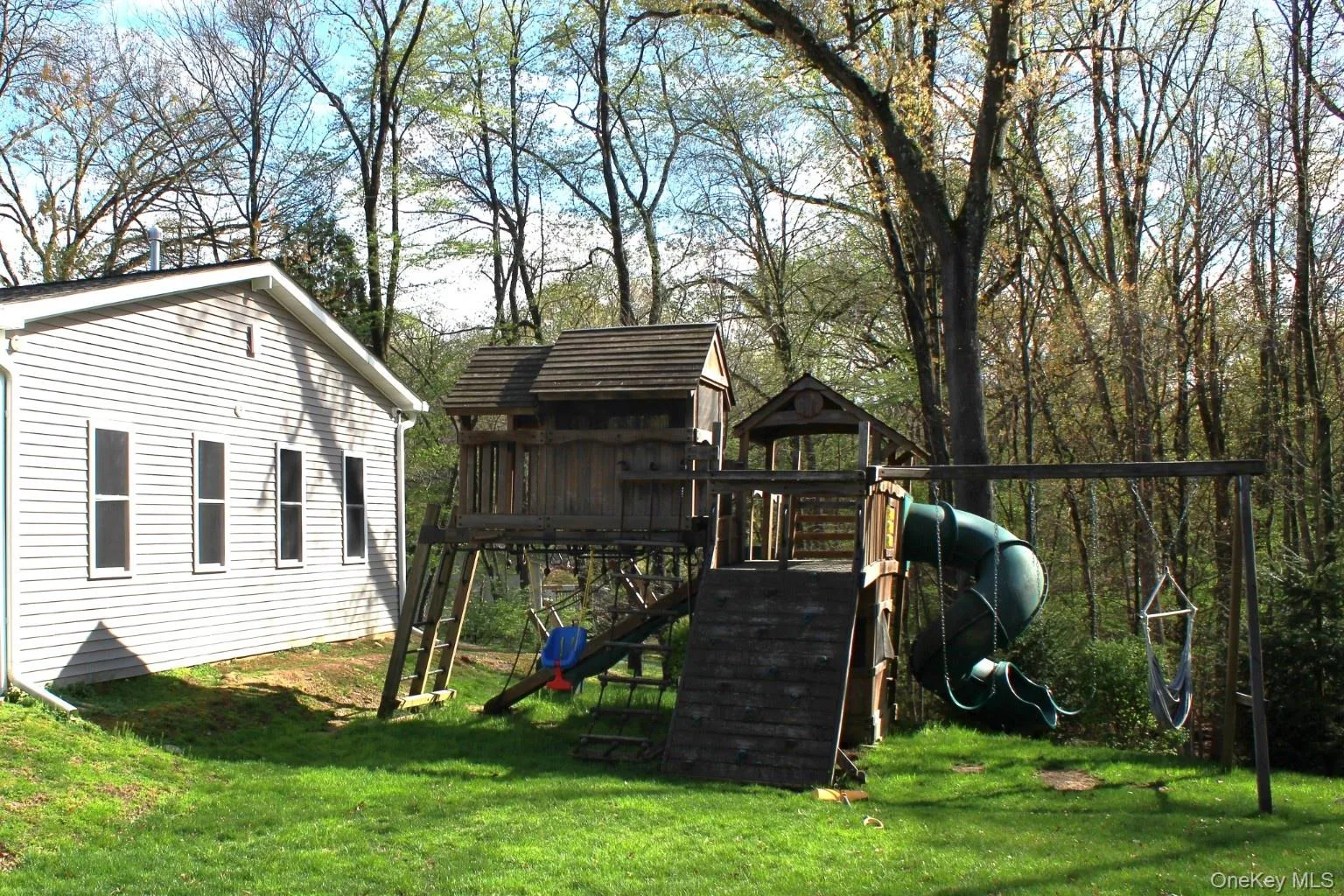 Expansive backyard featuring a lush lawn, mature trees, and a multi-level wooden play structure with a slide and swing set Expansive backyard featuring a lush lawn, mature trees, and a multi-level wooden play structure with a slide and swing set