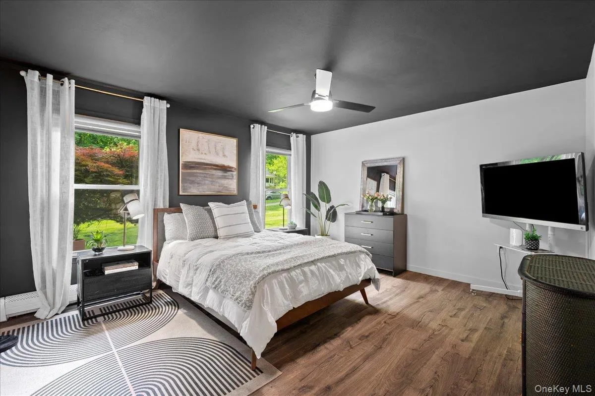 Bright and spacious main-floor bedroom with wood-finish flooring, two large windows, a ceiling fan with integrated lighting, and a calming neutral palette. Bright and spacious main-floor bedroom with wood-finish flooring, two large windows, a ceiling fan with integrated lighting, and a calming neutral palette.