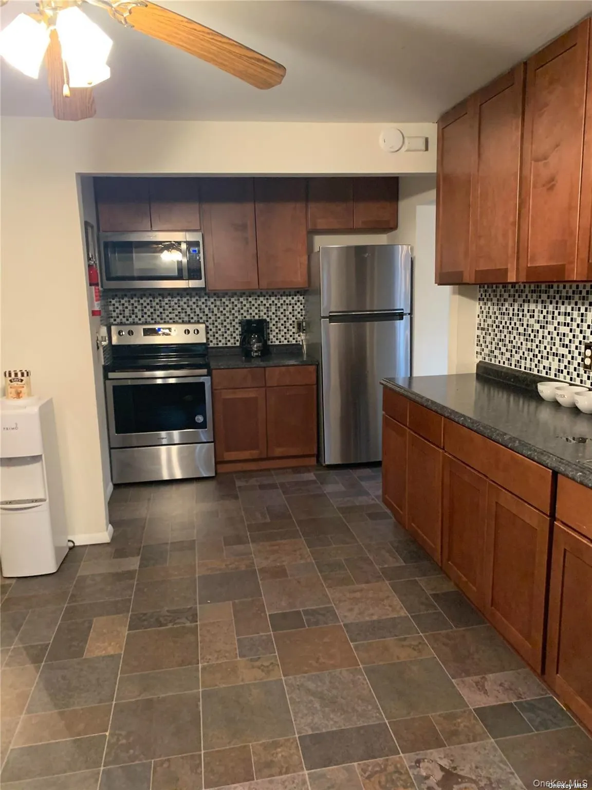 Kitchen with brown cabinets, ceiling fan, backsplash, dark countertops, and appliances with stainless steel finishes Kitchen with brown cabinets, ceiling fan, backsplash, dark countertops, and appliances with stainless steel finishes