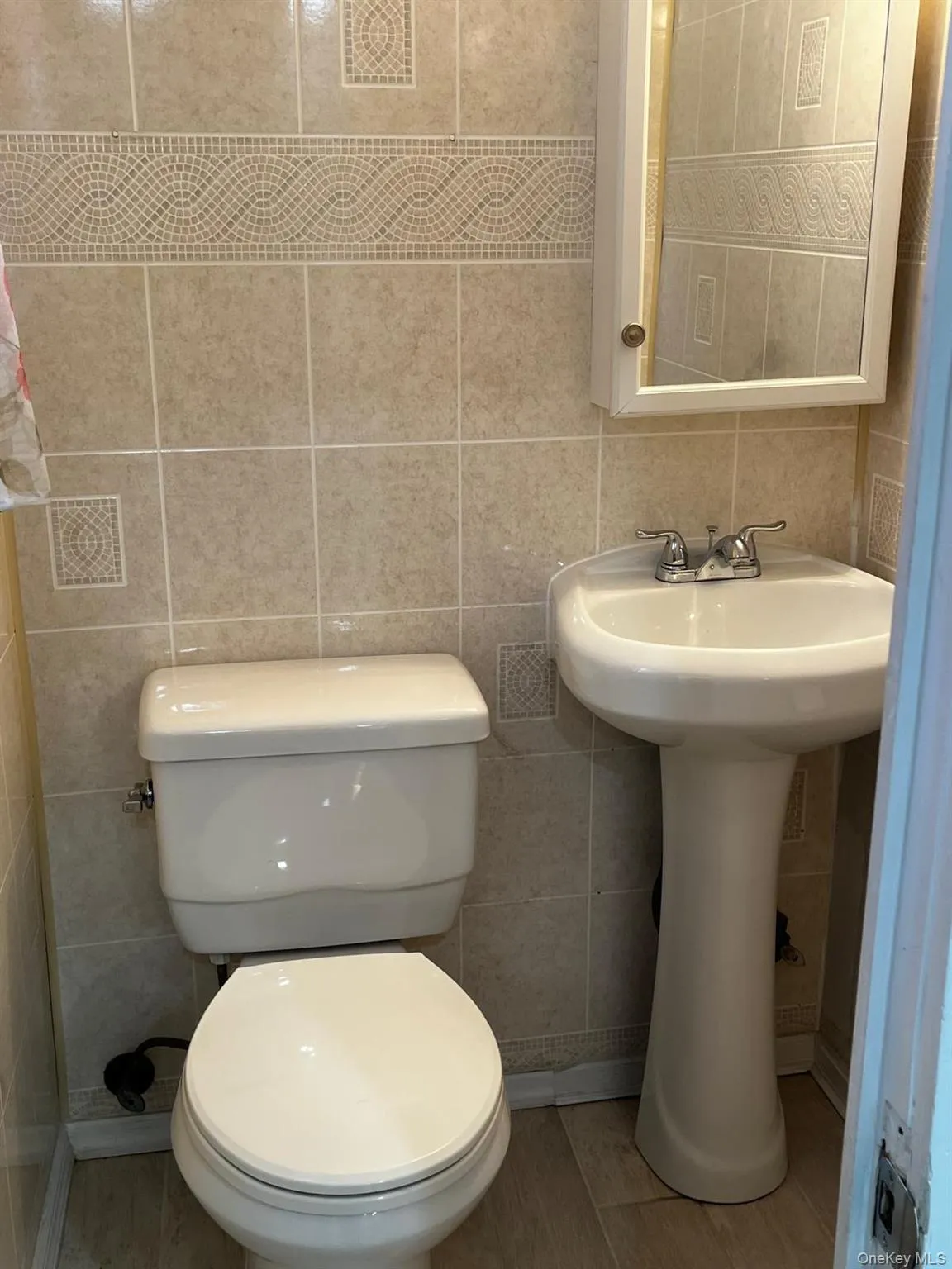 Bathroom featuring floor-to-ceiling ceramic tile, a decorative tile border, pedestal sink with chrome faucet, toilet, and a medicine cabinet with integrated mirror Bathroom featuring floor-to-ceiling ceramic tile, a decorative tile border, pedestal sink with chrome faucet, toilet, and a medicine cabinet with integrated mirror