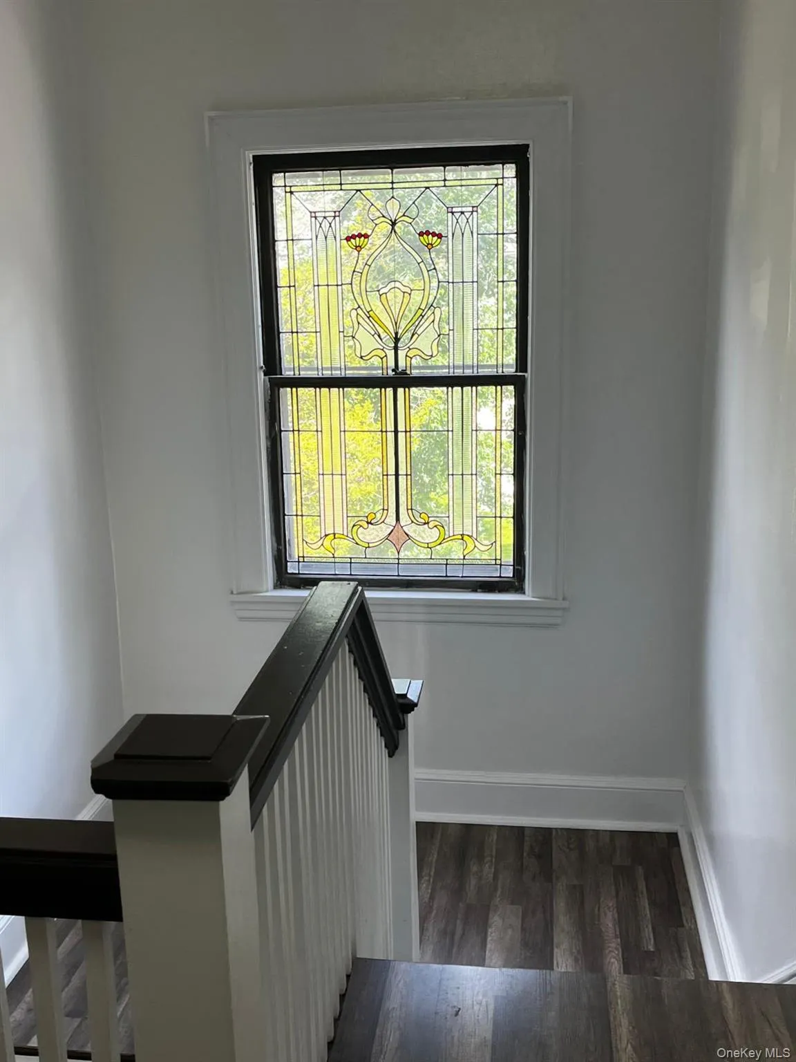 Staircase landing featuring a decorative stained glass window, dark wood-finish flooring, white painted walls, and a white and dark-stained wood balustrade Staircase landing featuring a decorative stained glass window, dark wood-finish flooring, white painted walls, and a white and dark-stained wood balustrade