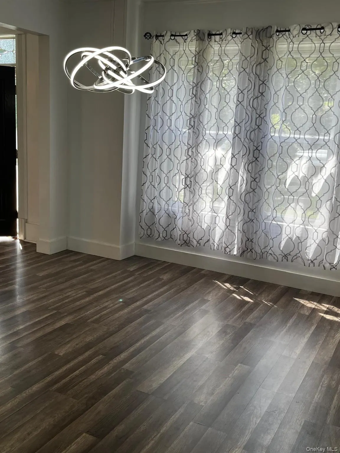 Wood-finish flooring with baseboard trim, contemporary light fixture, and window with sheer patterned curtains Wood-finish flooring with baseboard trim, contemporary light fixture, and window with sheer patterned curtains