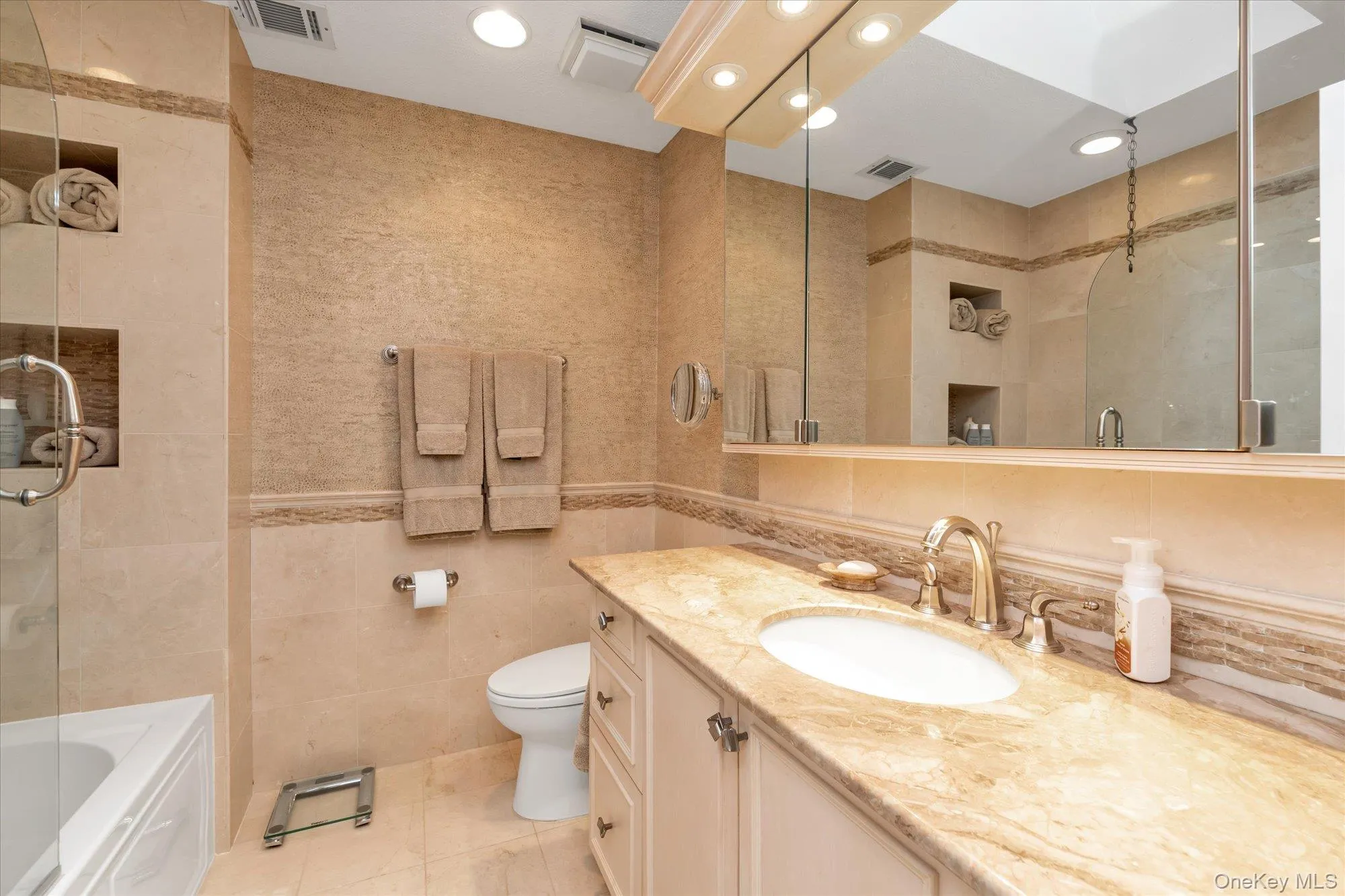 Bathroom featuring a vanity with an integrated sink and marble countertop, recessed lighting, a built-in shower/tub combination with a glass enclosure, and wall-mounted storage niches Bathroom featuring a vanity with an integrated sink and marble countertop, recessed lighting, a built-in shower/tub combination with a glass enclosure, and wall-mounted storage niches