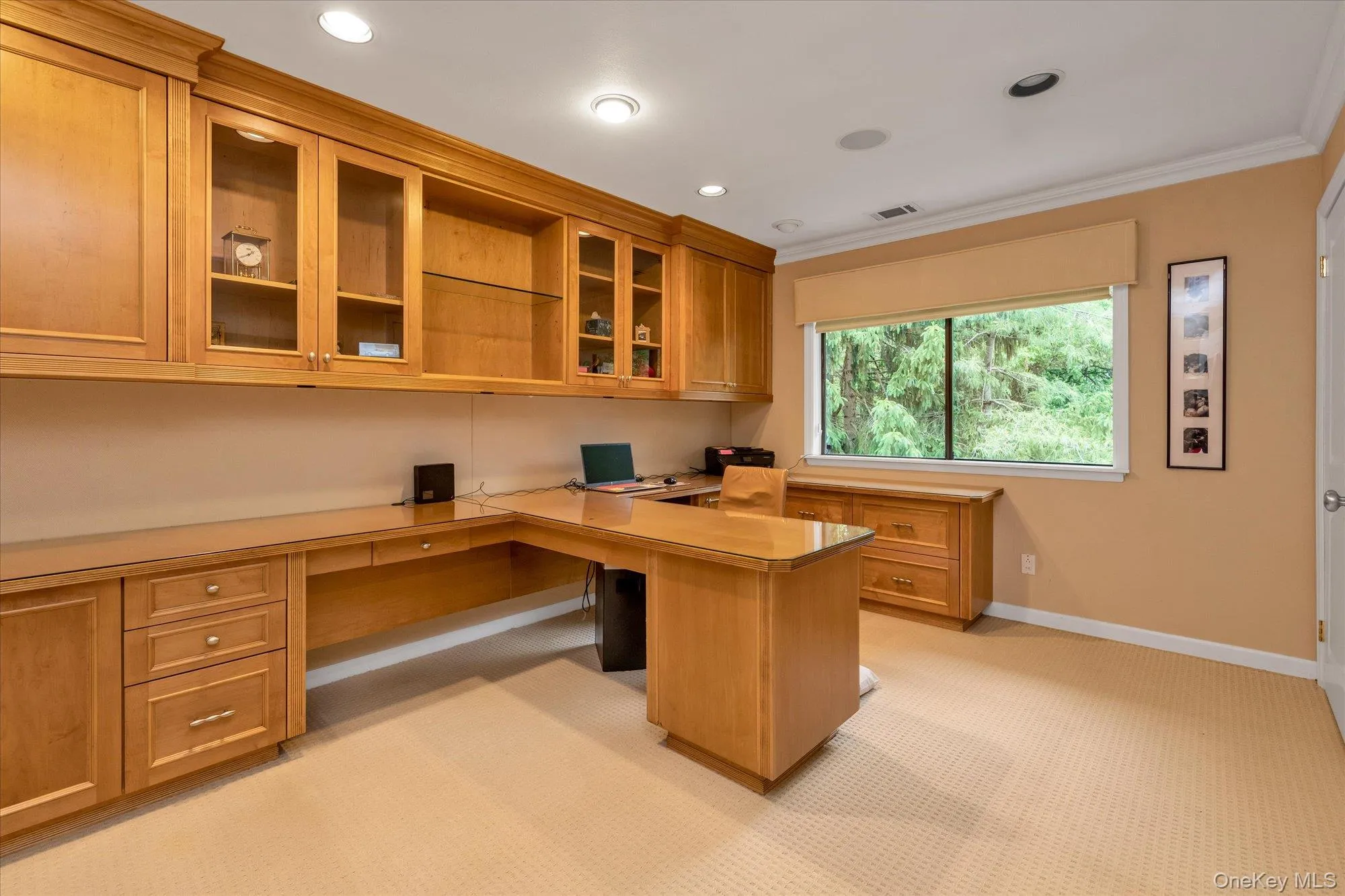 Bedroom with built-in wood cabinetry with glass-front uppers, L-shaped desk, recessed lighting, crown molding, and a large window overlooking mature trees Bedroom with built-in wood cabinetry with glass-front uppers, L-shaped desk, recessed lighting, crown molding, and a large window overlooking mature trees