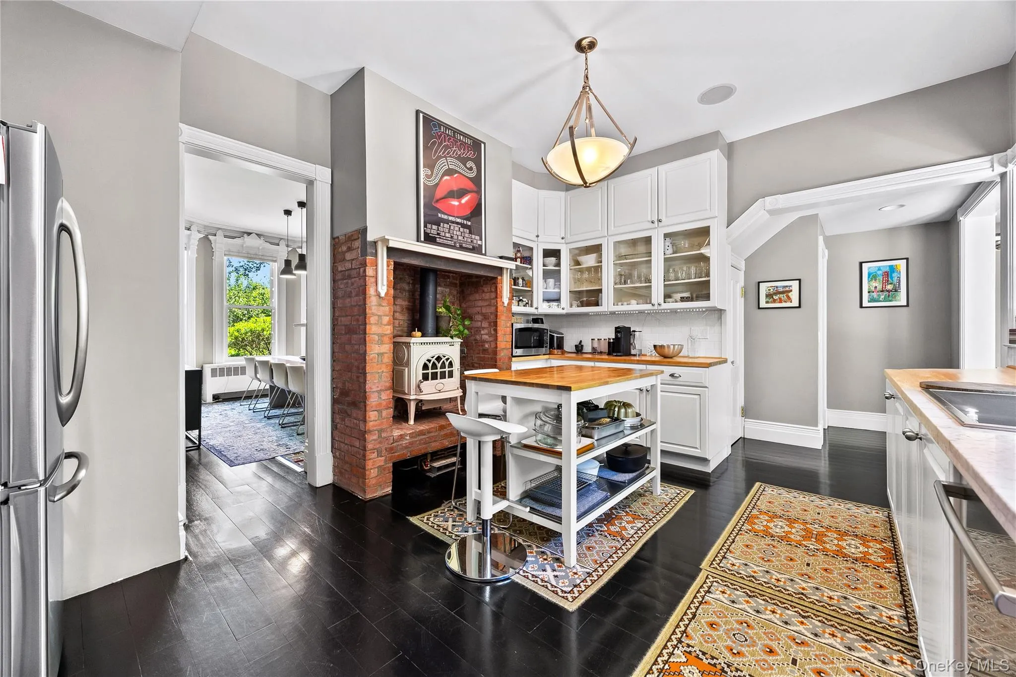 Kitchen featuring dark wood-finish flooring, white cabinetry with glass-front uppers, wood countertops, a brick-faced fireplace with a wood-burning stove, and a central island with integrated shelving Kitchen featuring dark wood-finish flooring, white cabinetry with glass-front uppers, wood countertops, a brick-faced fireplace with a wood-burning stove, and a central island with integrated shelving