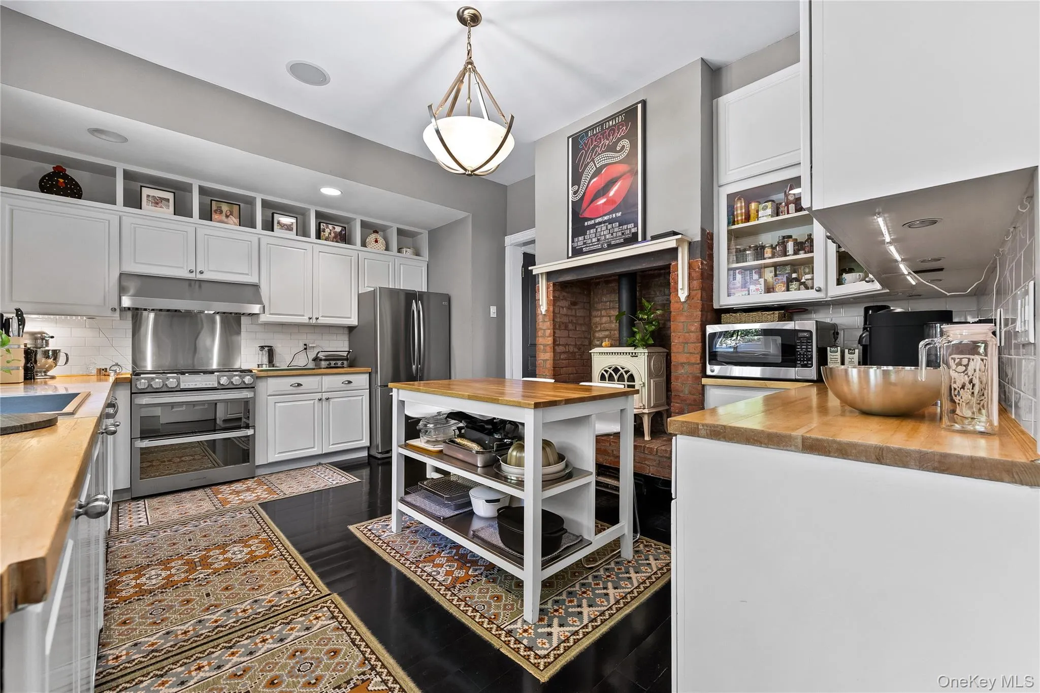 Gourmet kitchen featuring a stainless steel gas range, coordinating refrigerator, white shaker cabinetry with open shelving, wood-finish countertops, and a brick-surround fireplace Gourmet kitchen featuring a stainless steel gas range, coordinating refrigerator, white shaker cabinetry with open shelving, wood-finish countertops, and a brick-surround fireplace