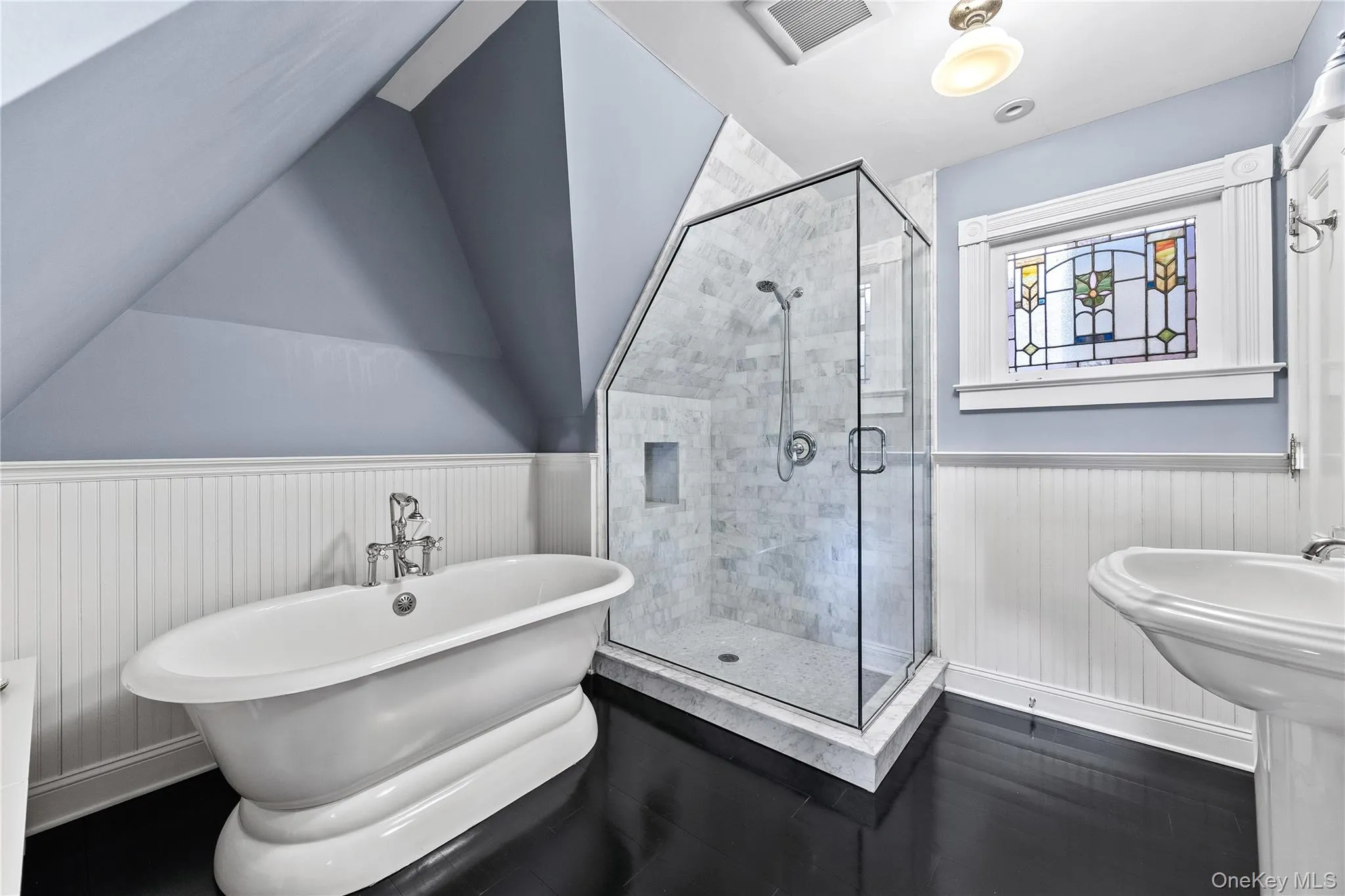 Bathroom featuring a freestanding bathtub, glass-enclosed shower with tiled surround, pedestal sink, dark wood-finish flooring, and a decorative stained-glass window Bathroom featuring a freestanding bathtub, glass-enclosed shower with tiled surround, pedestal sink, dark wood-finish flooring, and a decorative stained-glass window