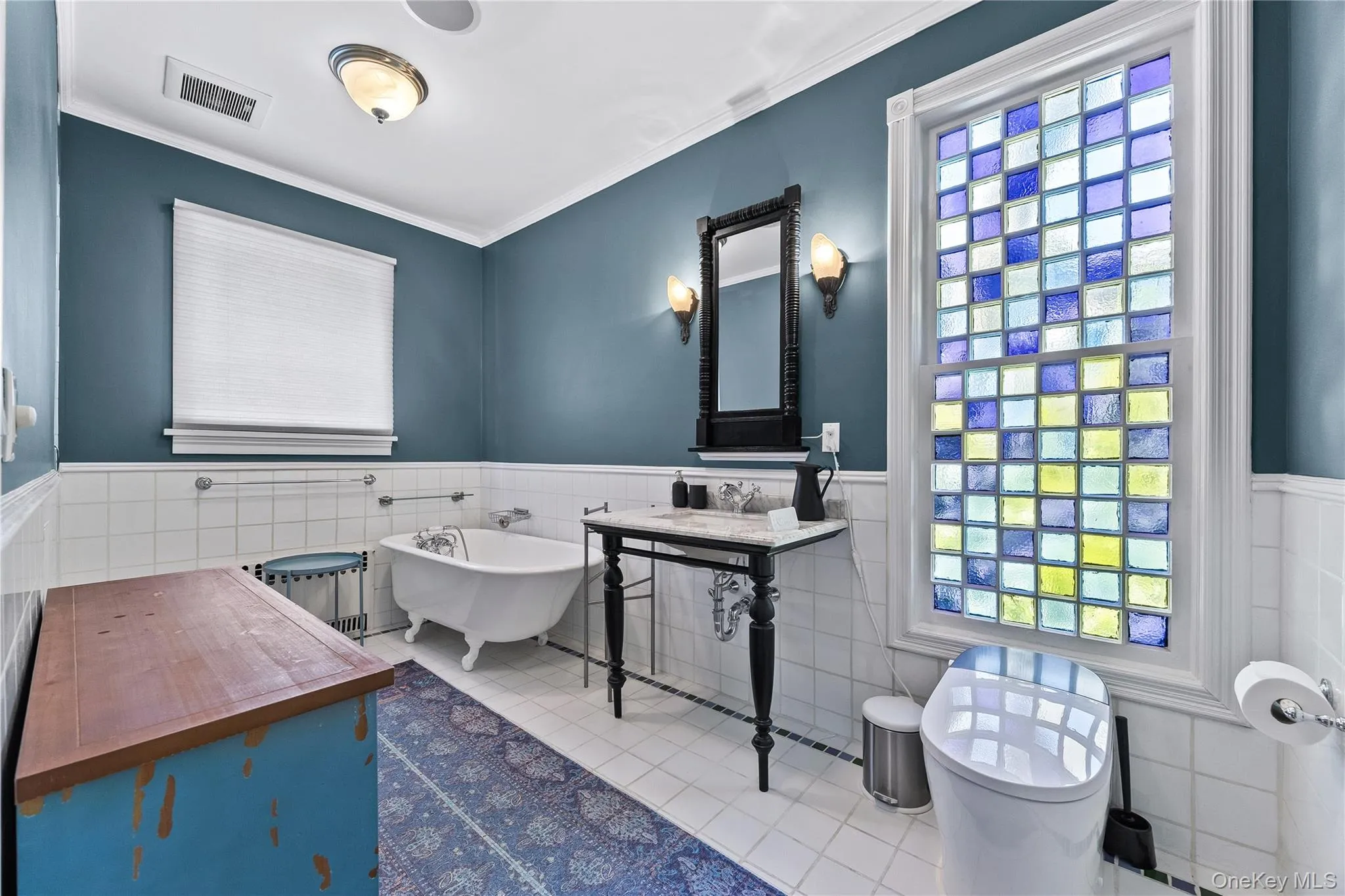 Bathroom featuring a colorful glass block window, clawfoot tub, white tile flooring, and a pedestal vanity with a marble top Bathroom featuring a colorful glass block window, clawfoot tub, white tile flooring, and a pedestal vanity with a marble top