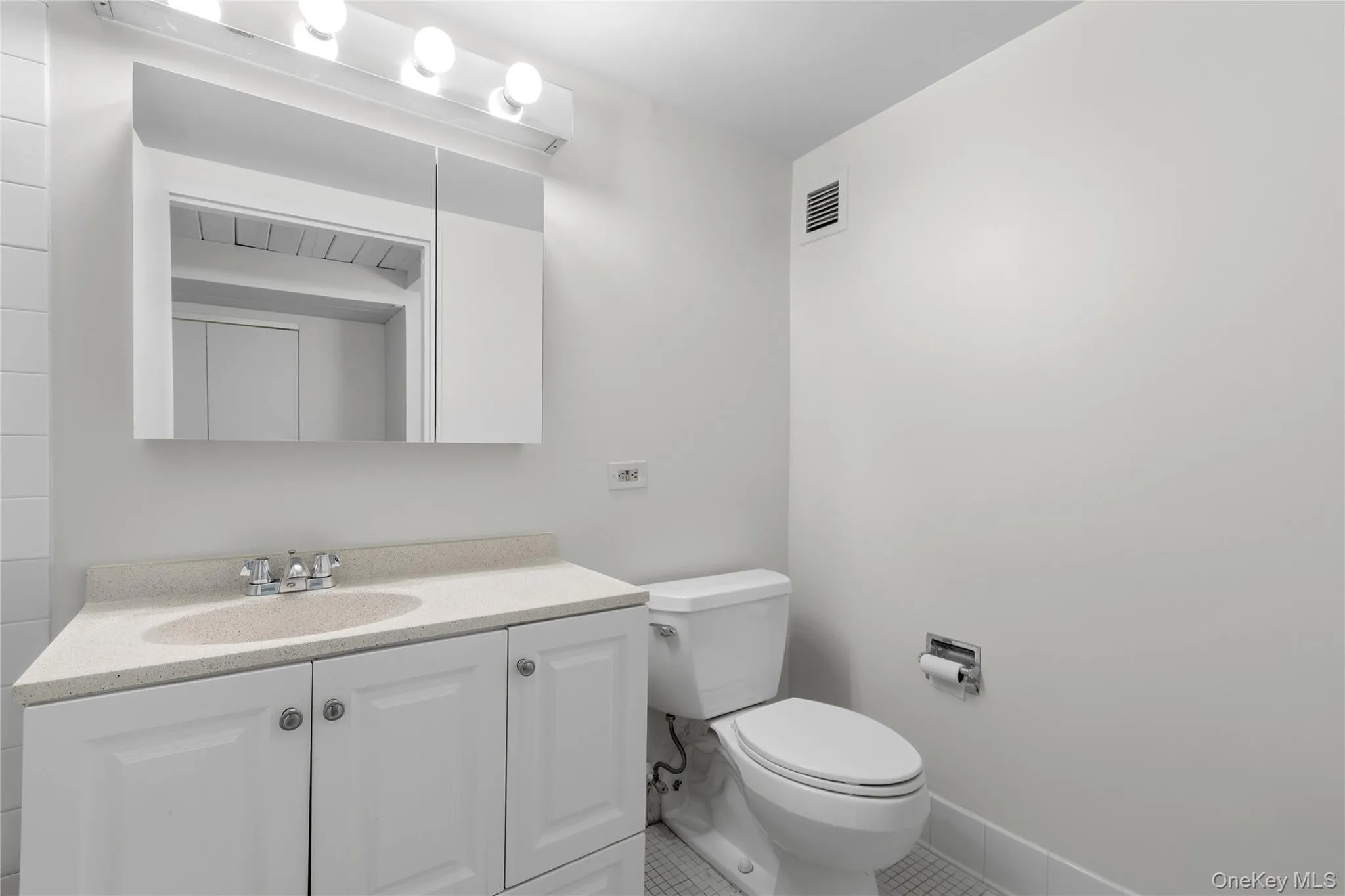 Bathroom featuring a white vanity with panel cabinetry, a light-toned countertop with an integrated sink, a mirrored medicine cabinet, and a white toilet Bathroom featuring a white vanity with panel cabinetry, a light-toned countertop with an integrated sink, a mirrored medicine cabinet, and a white toilet