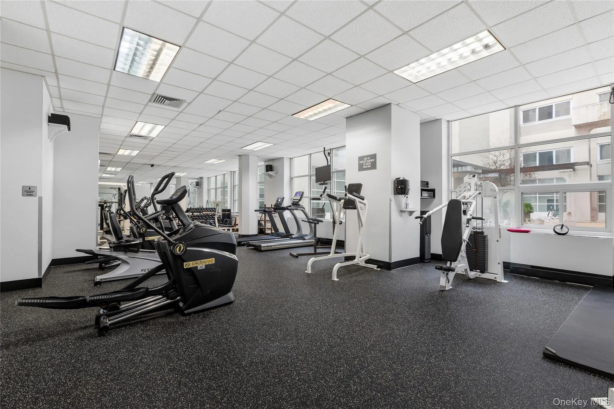 Dedicated fitness center featuring cardio and weight training equipment, resilient flooring, extensive window-walls, and recessed ceiling lighting Dedicated fitness center featuring cardio and weight training equipment, resilient flooring, extensive window-walls, and recessed ceiling lighting