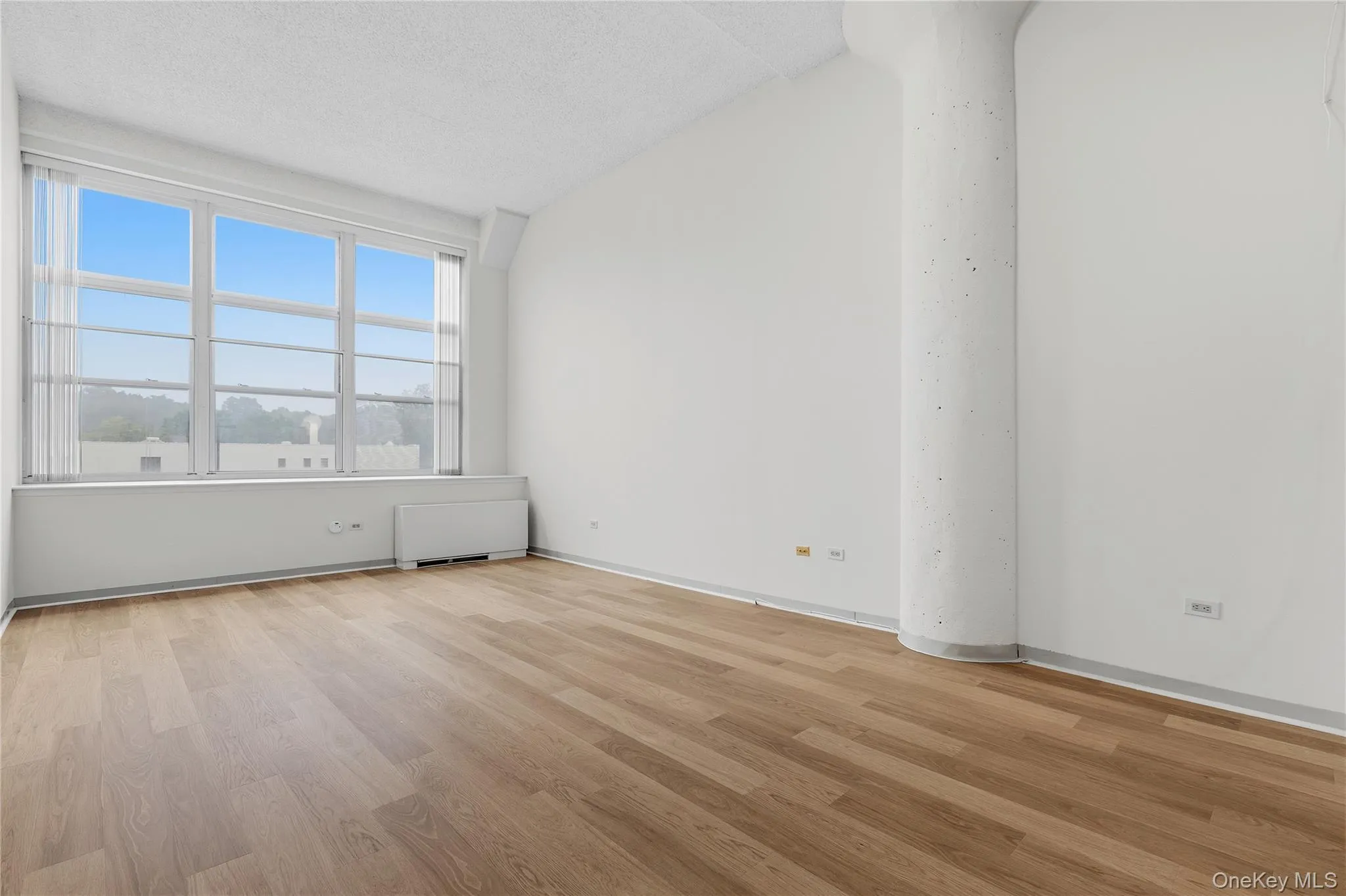 Open-concept room featuring wood-finish flooring, a full-wall window with vertical blinds, and a textured concrete column Open-concept room featuring wood-finish flooring, a full-wall window with vertical blinds, and a textured concrete column