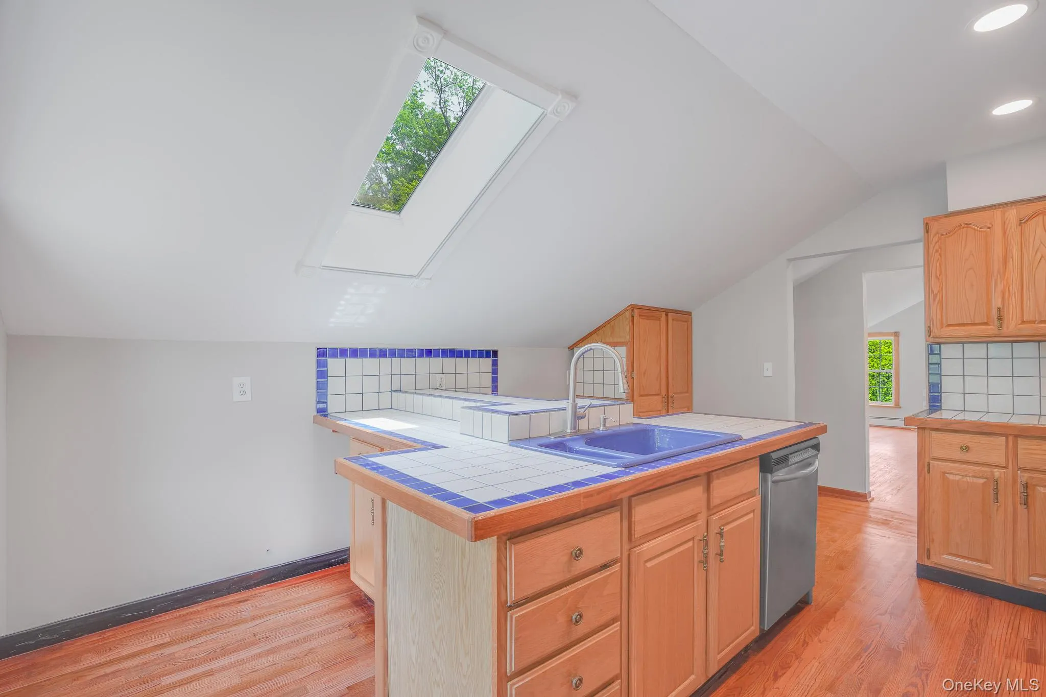 Kitchen featuring a skylight, wood-finish flooring, wood cabinetry, tile countertops with a blue trim, and a built-in dishwasher Kitchen featuring a skylight, wood-finish flooring, wood cabinetry, tile countertops with a blue trim, and a built-in dishwasher