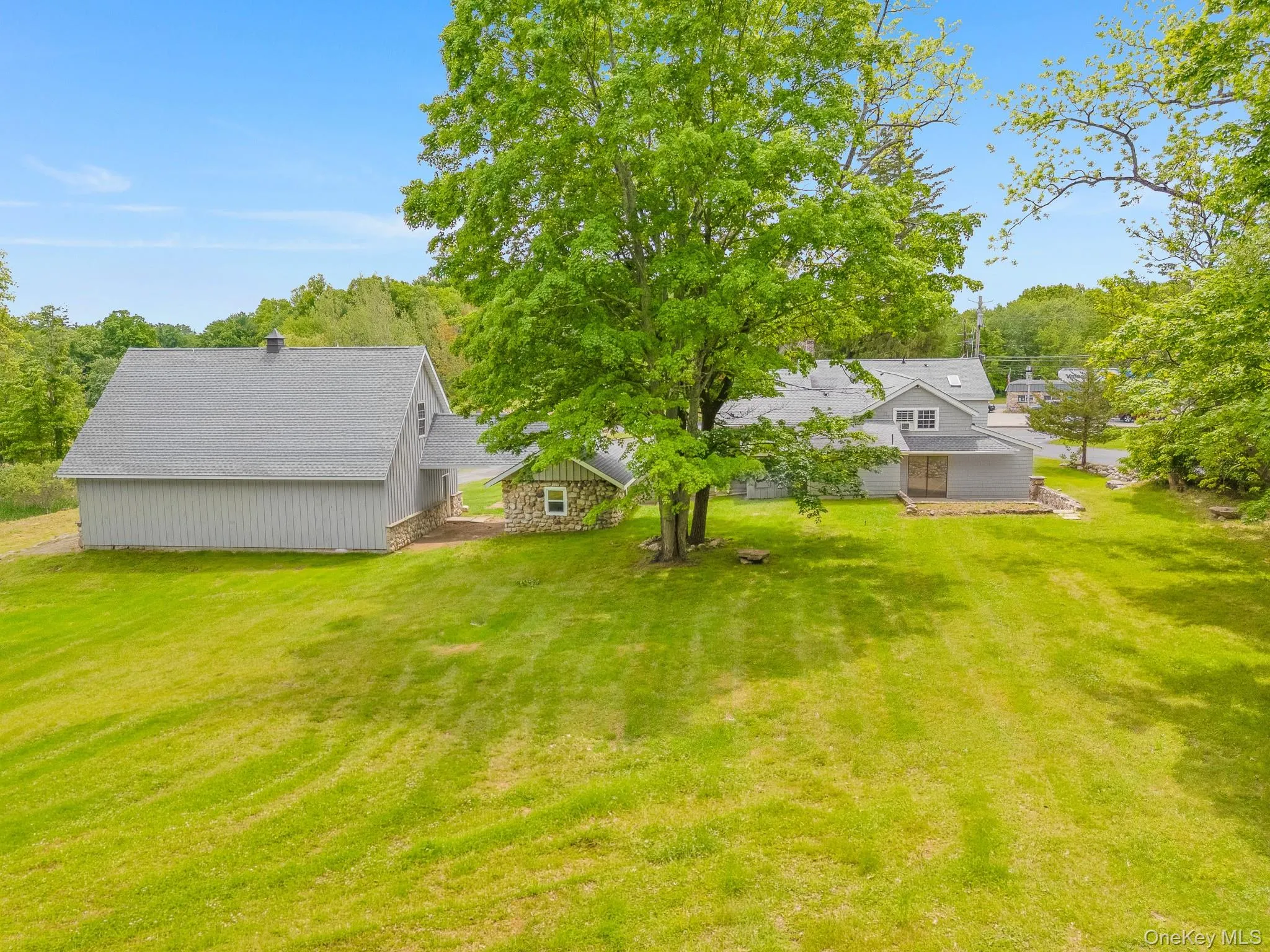 Expansive grassy yard area featuring multiple structures, including a large outbuilding with a gray shingle roof and vertical siding, a stone-clad shed, and a multi-level residence with a deck Expansive grassy yard area featuring multiple structures, including a large outbuilding with a gray shingle roof and vertical siding, a stone-clad shed, and a multi-level residence with a deck