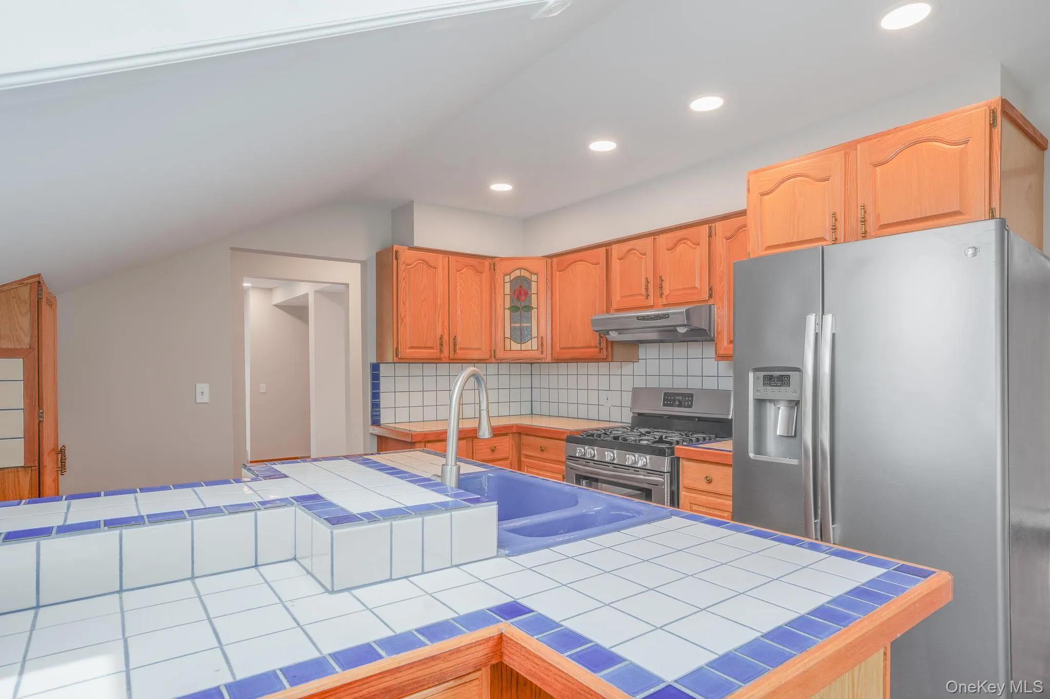 Kitchen featuring a tiled island with an integrated sink, wood-finish cabinetry, stainless steel appliances, a gas range with a vent hood, and recessed lighting Kitchen featuring a tiled island with an integrated sink, wood-finish cabinetry, stainless steel appliances, a gas range with a vent hood, and recessed lighting