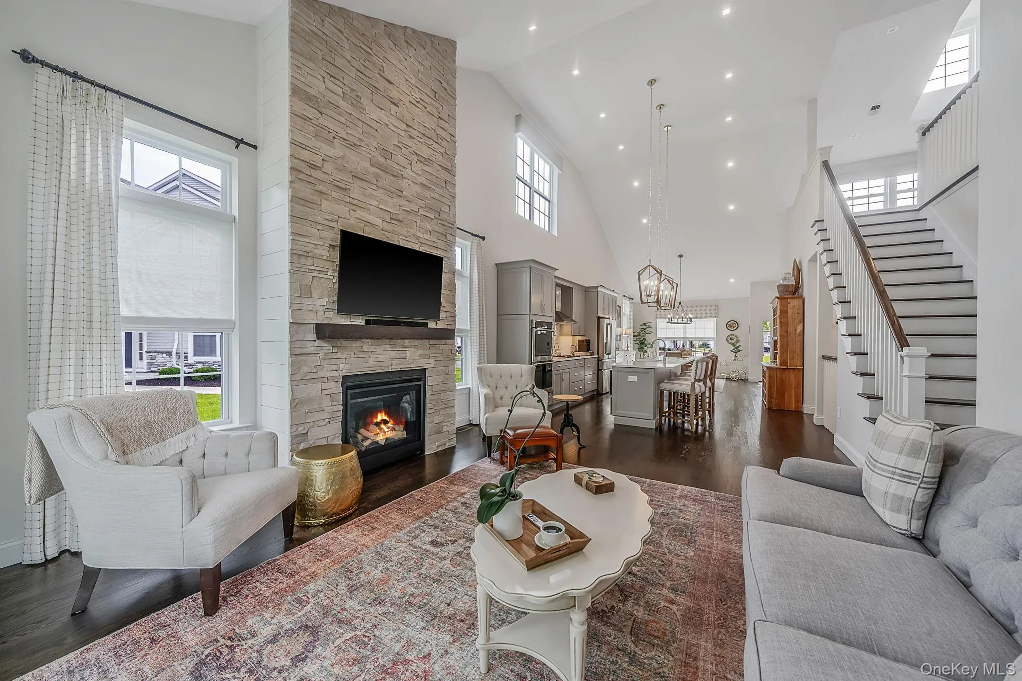Vaulted great room featuring a stone-clad fireplace with mantel, dark wood-finish flooring, recessed lighting, and a white staircase with dark wood-finish handrails Vaulted great room featuring a stone-clad fireplace with mantel, dark wood-finish flooring, recessed lighting, and a white staircase with dark wood-finish handrails