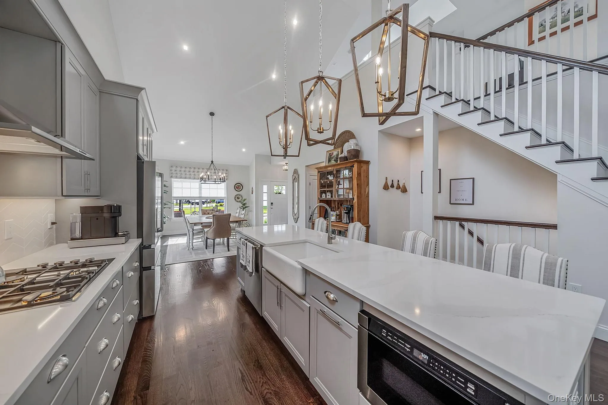 Open-concept kitchen featuring an oversized island with an integrated farmhouse sink, wood-finish flooring, light gray cabinetry, and white countertops Open-concept kitchen featuring an oversized island with an integrated farmhouse sink, wood-finish flooring, light gray cabinetry, and white countertops