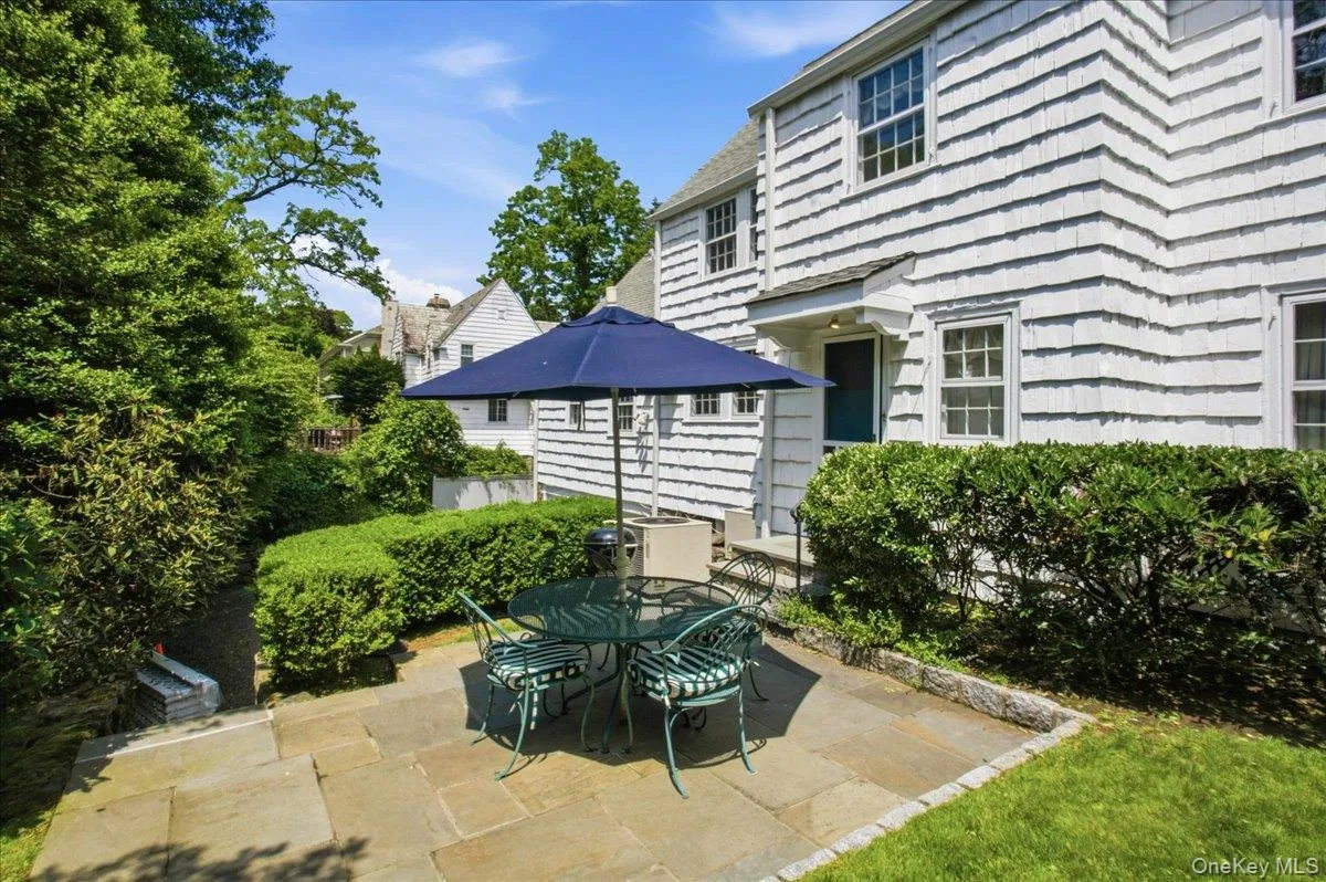 Stone patio with layered landscaping, white cedar shingle siding, a covered entry, and gridded windows Stone patio with layered landscaping, white cedar shingle siding, a covered entry, and gridded windows
