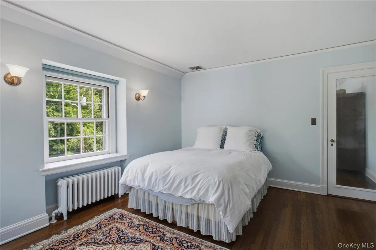 Hardwood flooring throughout this room features a large window with white muntins, crown molding, and two wall-mounted sconces Hardwood flooring throughout this room features a large window with white muntins, crown molding, and two wall-mounted sconces