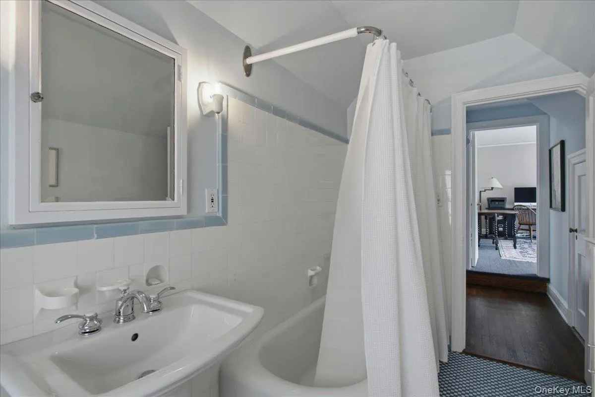 Hall bathroom, second floor, featuring a white pedestal sink with chrome fixtures, a medicine cabinet with integrated lighting, and white subway tile with a light blue accent border Hall bathroom, second floor, featuring a white pedestal sink with chrome fixtures, a medicine cabinet with integrated lighting, and white subway tile with a light blue accent border