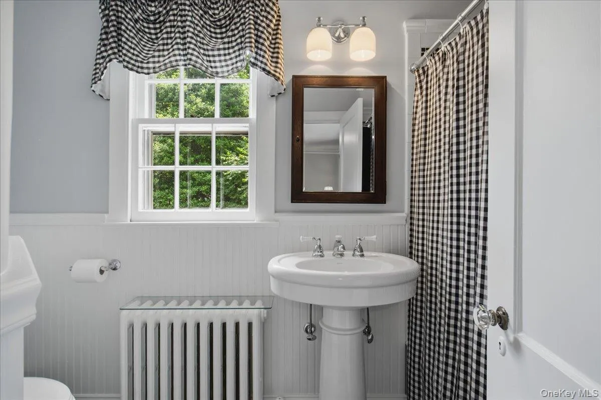 Pedestal sink with clear knob faucet handles, wood-framed mirror, two-light vanity fixture, white beadboard wainscoting, and a multi-pane window Pedestal sink with clear knob faucet handles, wood-framed mirror, two-light vanity fixture, white beadboard wainscoting, and a multi-pane window