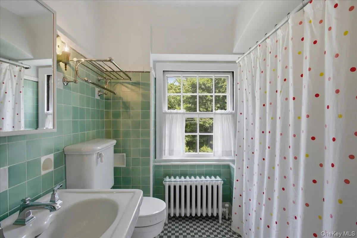 Bathroom featuring green wall tiling, black and white checkered floor tiling, a white window, a white radiator, and a wall-mounted shelving unit Bathroom featuring green wall tiling, black and white checkered floor tiling, a white window, a white radiator, and a wall-mounted shelving unit