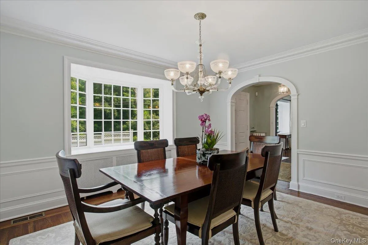 Dining area featuring a bay window with grid-patterned panes, an ornate brass chandelier, elegant crown molding, decorative wainscoting, and a wide arched entryway Dining area featuring a bay window with grid-patterned panes, an ornate brass chandelier, elegant crown molding, decorative wainscoting, and a wide arched entryway