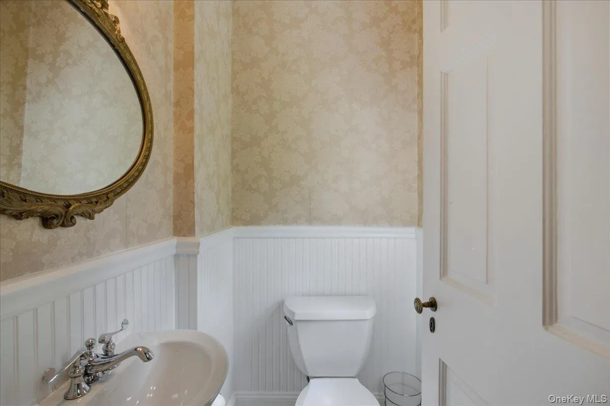 First floor powder room featuring a pedestal sink with a chrome faucet, ornate framed mirror, wainscoting, and patterned wallpaper First floor powder room featuring a pedestal sink with a chrome faucet, ornate framed mirror, wainscoting, and patterned wallpaper