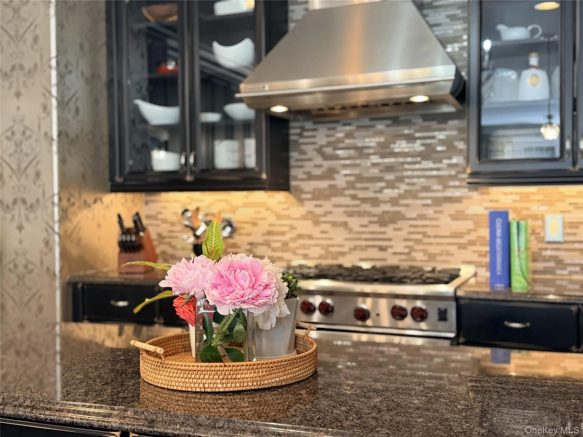 Kitchen featuring dark stone countertops, mosaic tile backsplash, professional-grade gas range, stainless steel range hood, and dark cabinetry with glass panel doors Kitchen featuring dark stone countertops, mosaic tile backsplash, professional-grade gas range, stainless steel range hood, and dark cabinetry with glass panel doors
