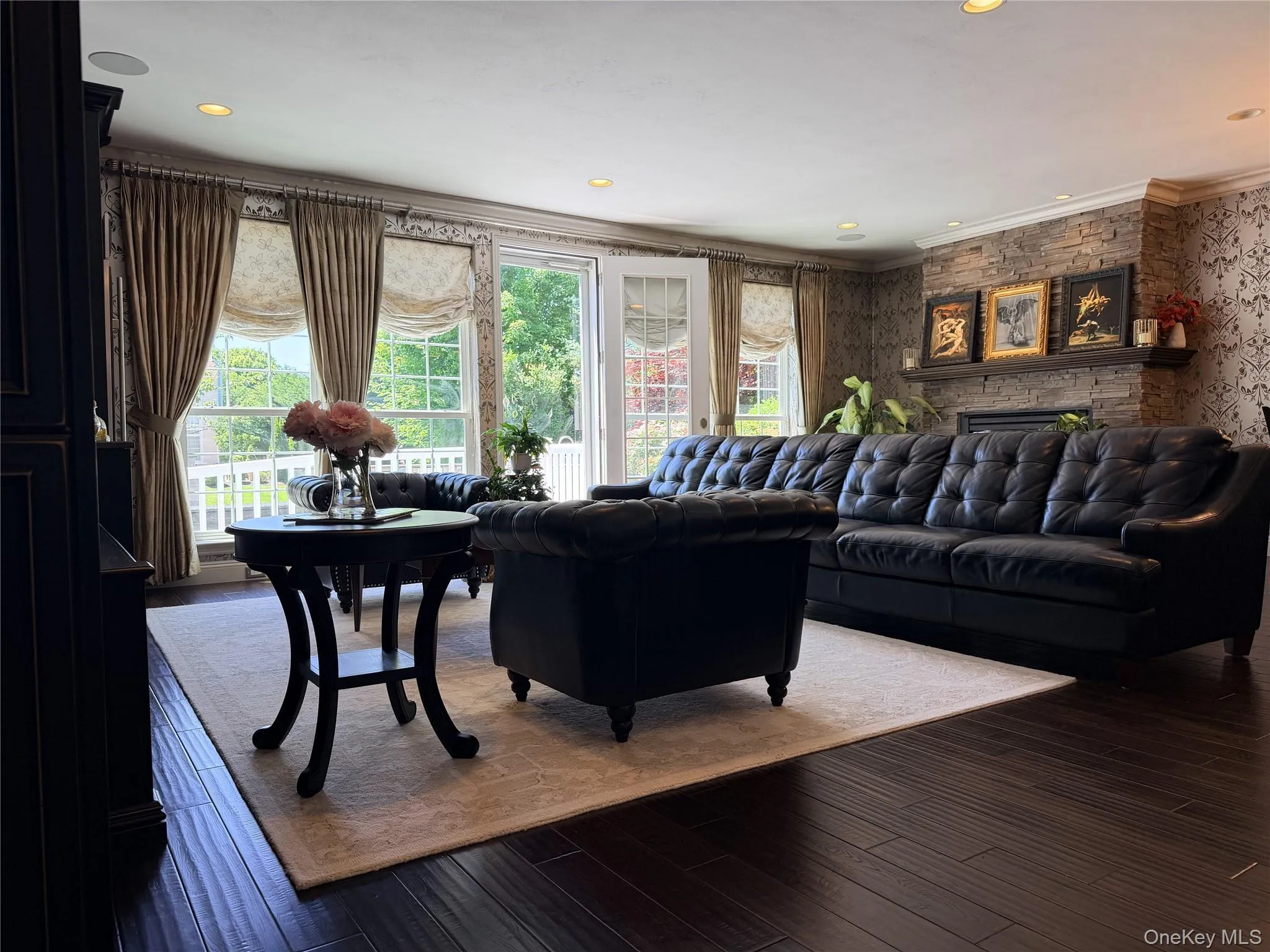 Living area featuring rich wood-finish flooring, a stone-faced fireplace with a mantel, recessed lighting, crown molding, and direct access to an exterior balcony Living area featuring rich wood-finish flooring, a stone-faced fireplace with a mantel, recessed lighting, crown molding, and direct access to an exterior balcony