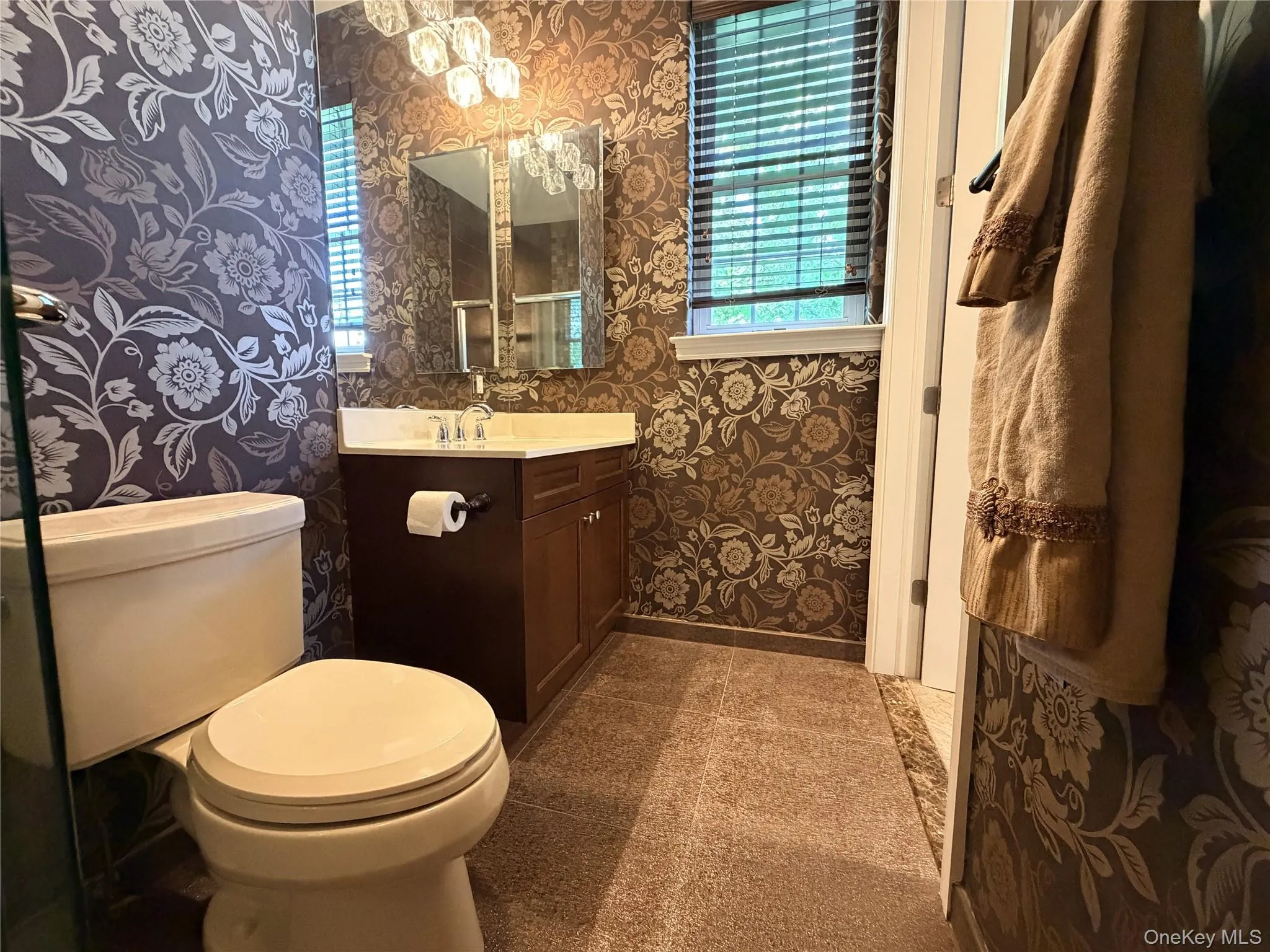 Bathroom featuring patterned wallpaper, a single vanity with a white countertop, a mirror, and a multi-bulb light fixture Bathroom featuring patterned wallpaper, a single vanity with a white countertop, a mirror, and a multi-bulb light fixture