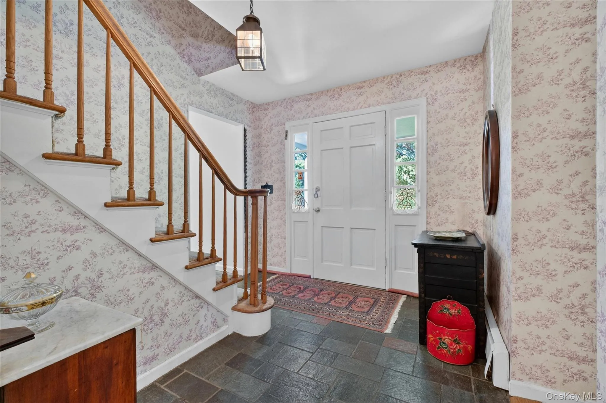 Foyer featuring raised panel front door with side light windows, dark slate tile flooring, a curved wood handrail, and patterned wallpaper Foyer featuring raised panel front door with side light windows, dark slate tile flooring, a curved wood handrail, and patterned wallpaper