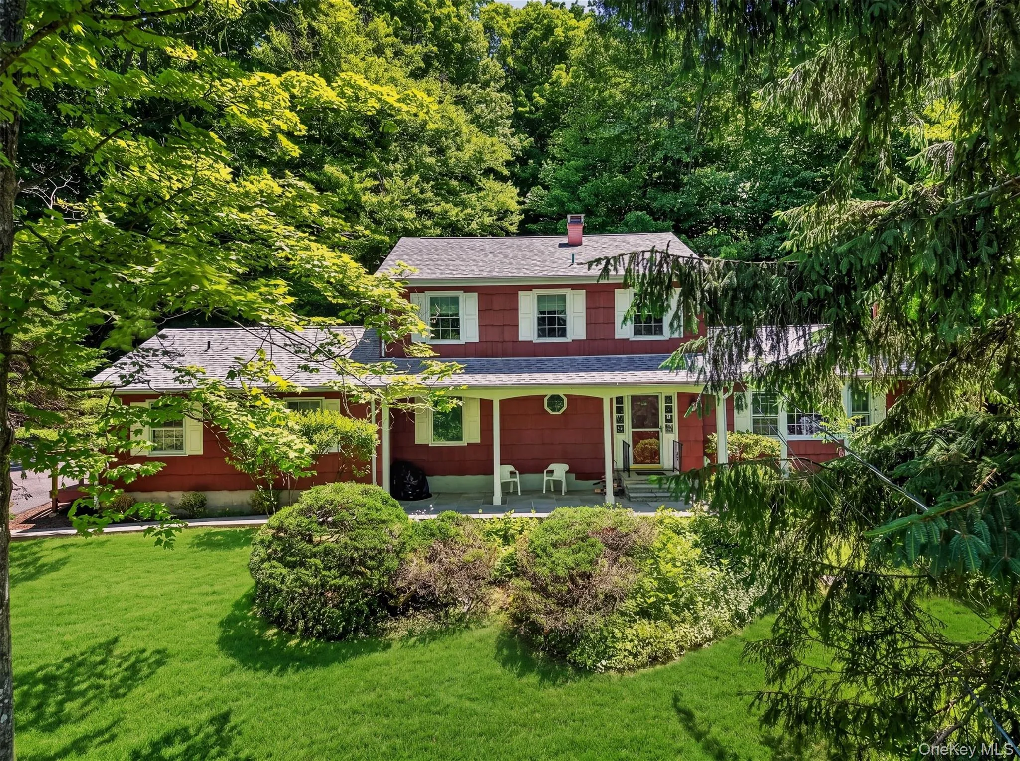 Residence featuring covered front porch, white window shutters, a grey shingled roof, and a verdant lawn with mature trees and shrubs Residence featuring covered front porch, white window shutters, a grey shingled roof, and a verdant lawn with mature trees and shrubs