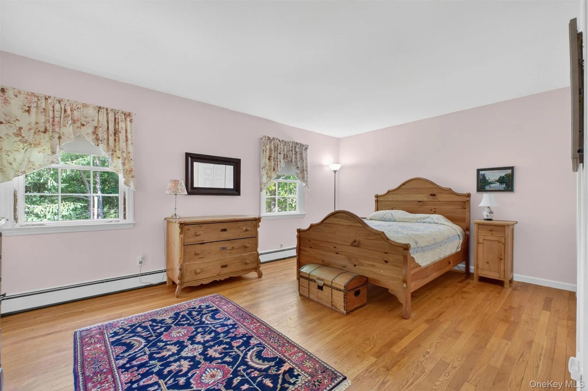 Primary Bedroom w/Hardwood flooring throughout featuring two windows with white trim and a baseboard heating system Primary Bedroom w/Hardwood flooring throughout featuring two windows with white trim and a baseboard heating system