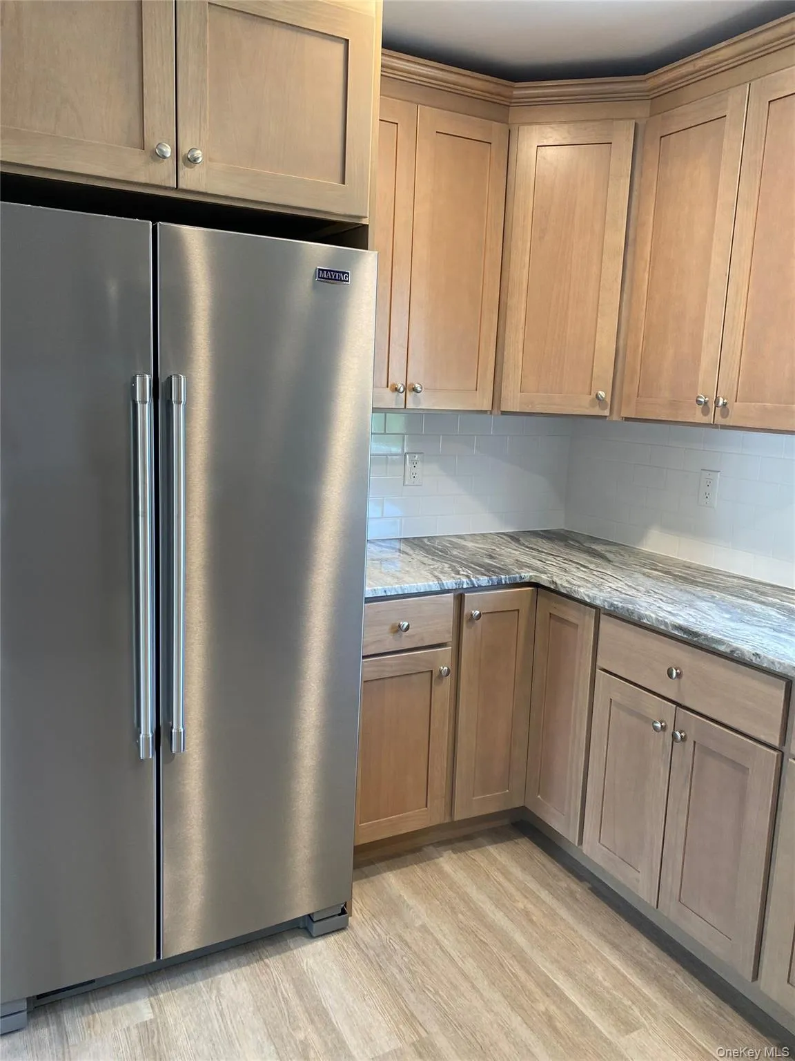 Kitchen featuring high end refrigerator, decorative backsplash, light stone countertops, light wood-type flooring, and brown cabinetry Kitchen featuring high end refrigerator, decorative backsplash, light stone countertops, light wood-type flooring, and brown cabinetry