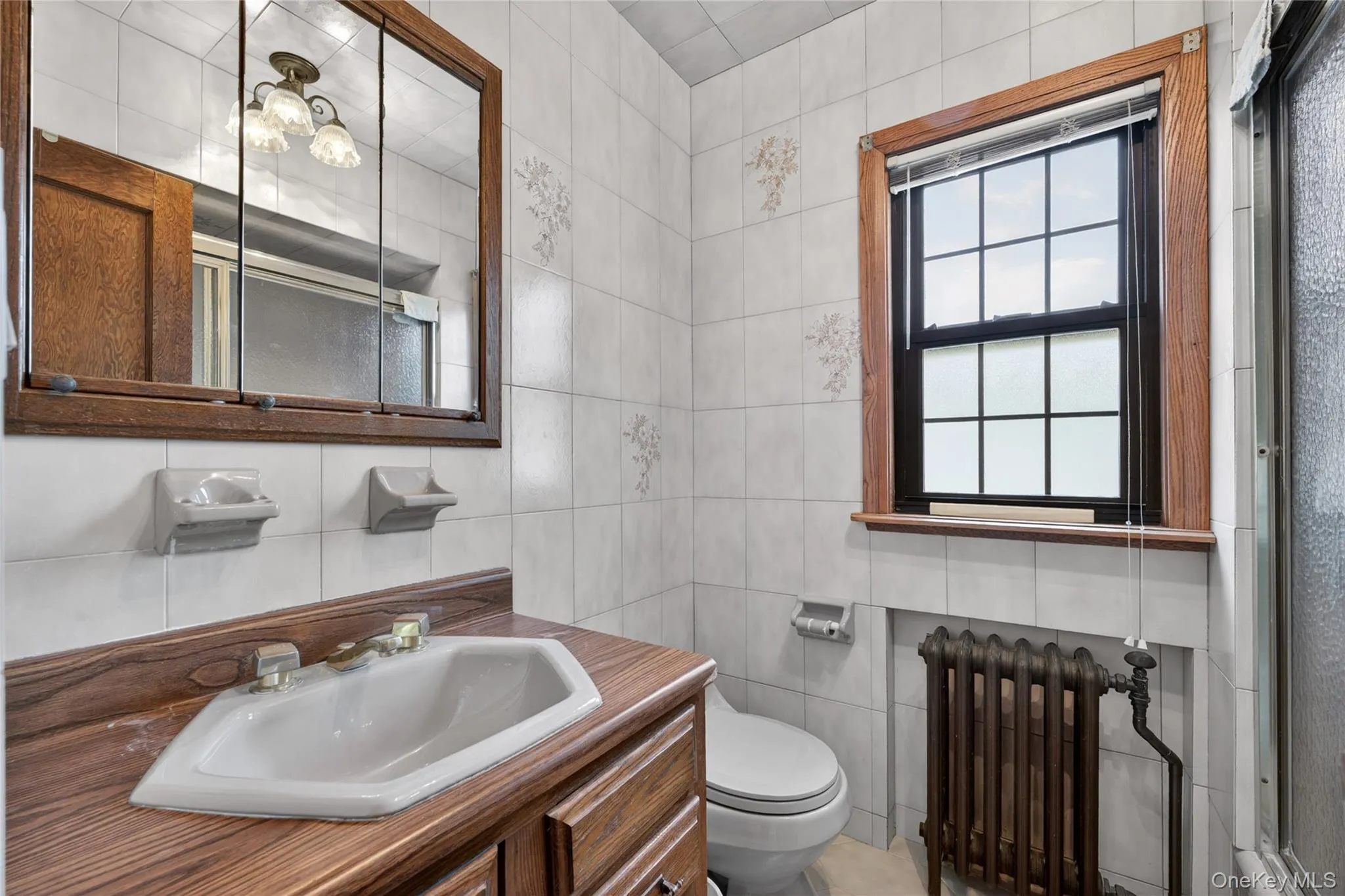Bathroom featuring ceramic tile walls with subtle floral accents, a wood-finish vanity with an integrated sink, a medicine cabinet with mirror panels, a window with a dark frame and light wood trim, and a cast iron radiator Bathroom featuring ceramic tile walls with subtle floral accents, a wood-finish vanity with an integrated sink, a medicine cabinet with mirror panels, a window with a dark frame and light wood trim, and a cast iron radiator