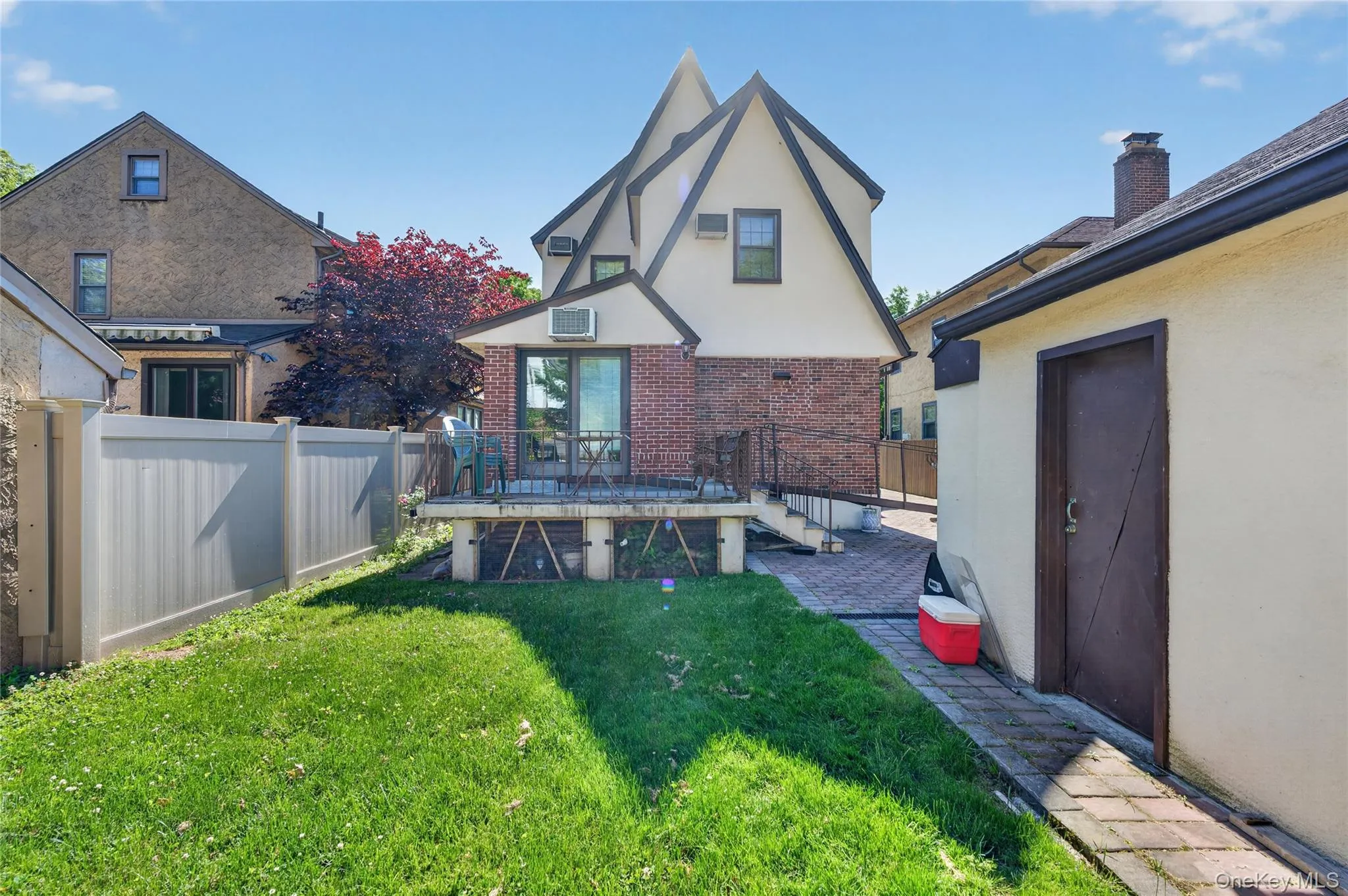 Rear exterior featuring a grass lawn, a small elevated deck with sliding glass doors, a brick patio, and a separate stucco-finished structure with a brown door Rear exterior featuring a grass lawn, a small elevated deck with sliding glass doors, a brick patio, and a separate stucco-finished structure with a brown door