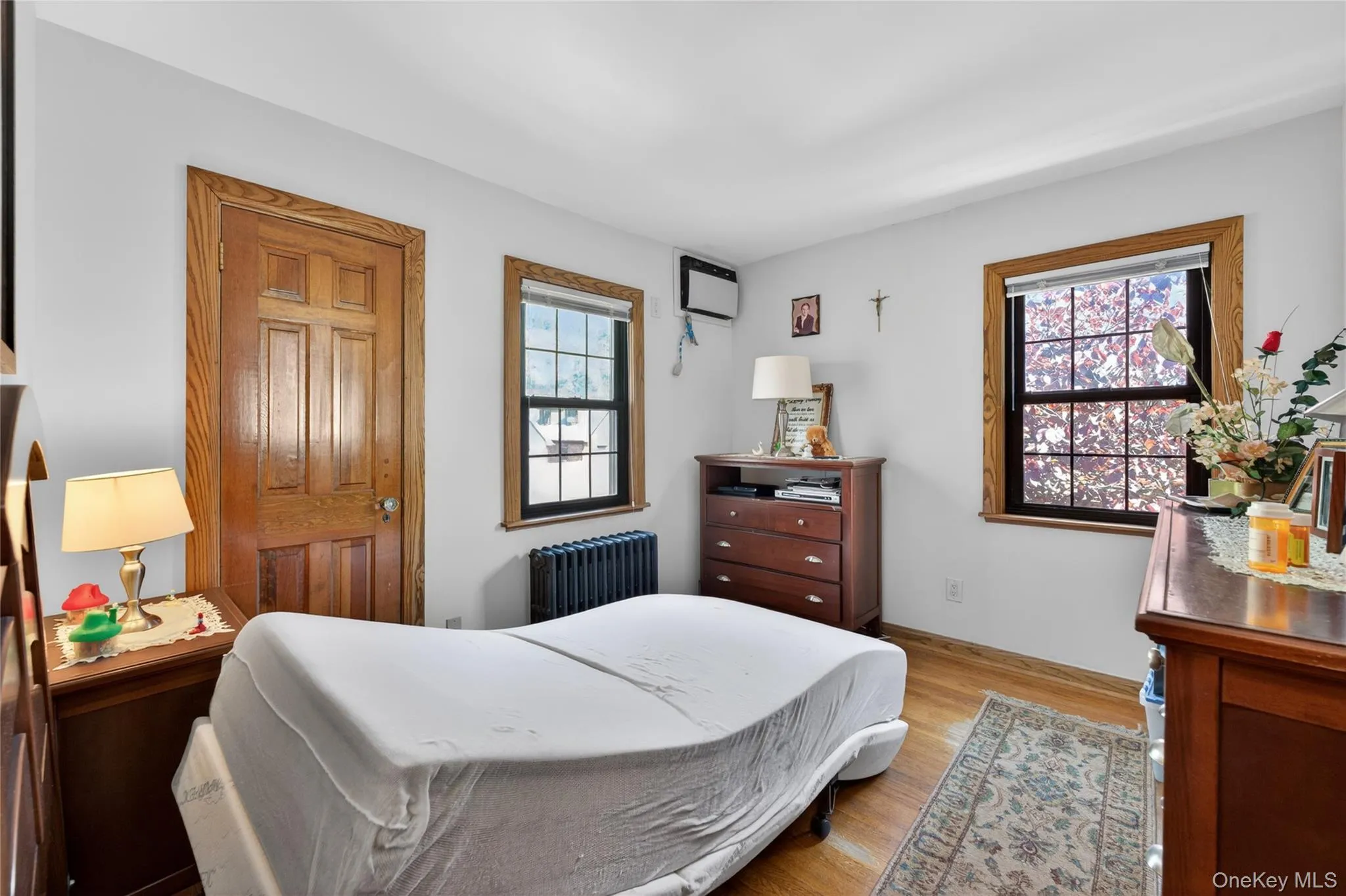 Room featuring wood-finish flooring, two windows with wood trim, a built-in radiator, a paneled wood door, and white walls Room featuring wood-finish flooring, two windows with wood trim, a built-in radiator, a paneled wood door, and white walls
