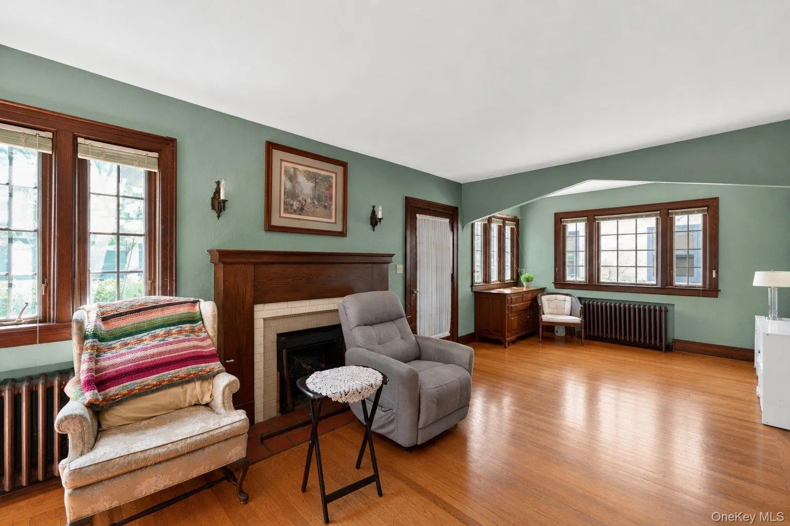Spacious living area featuring hardwood flooring, a prominent fireplace with a wood mantel, and multiple windows with dark wood trim Spacious living area featuring hardwood flooring, a prominent fireplace with a wood mantel, and multiple windows with dark wood trim