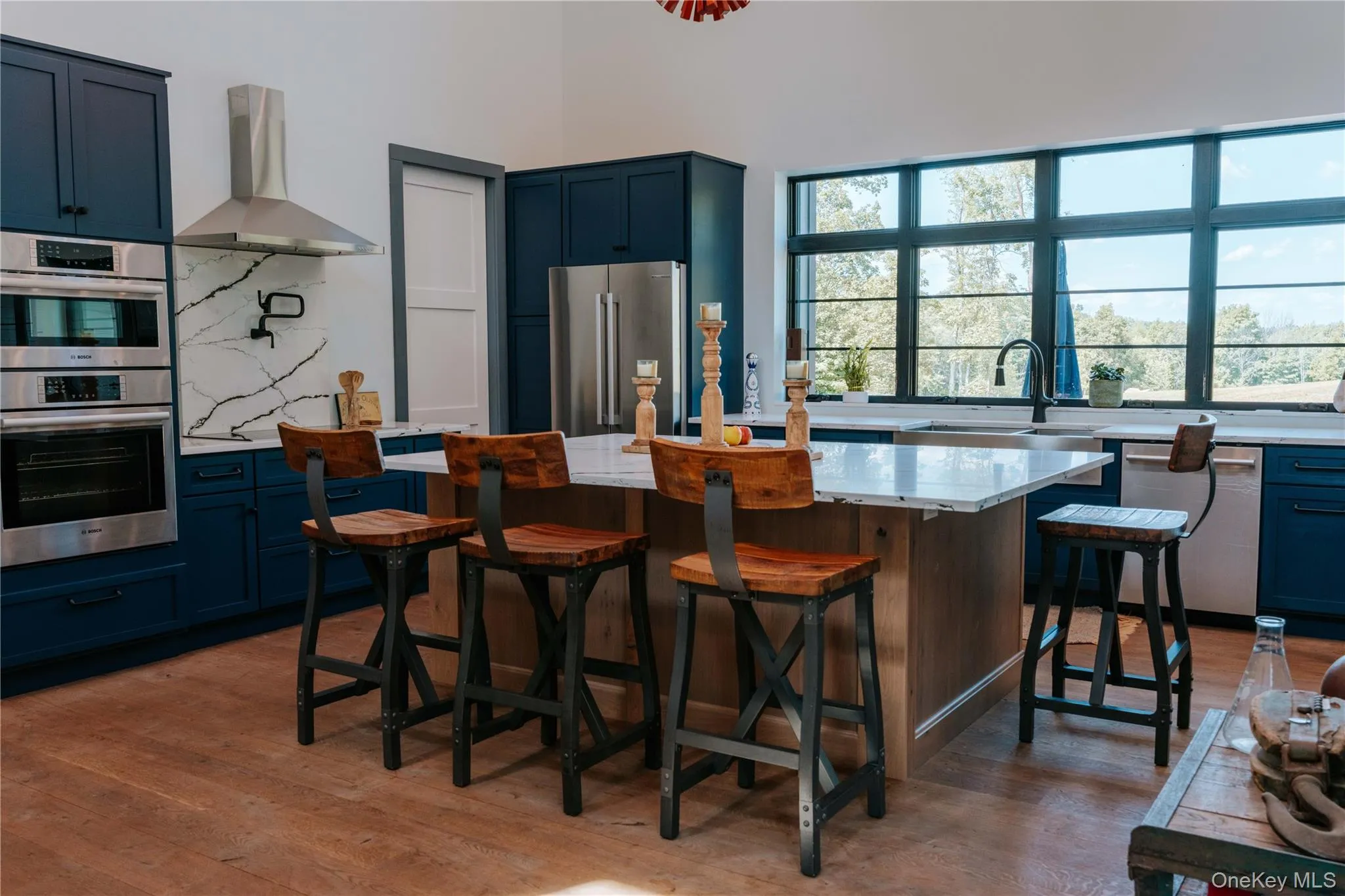 Kitchen with tasteful backsplash, blue cabinetry, wall chimney exhaust hood, appliances with stainless steel finishes, and light wood-type flooring Kitchen with tasteful backsplash, blue cabinetry, wall chimney exhaust hood, appliances with stainless steel finishes, and light wood-type flooring