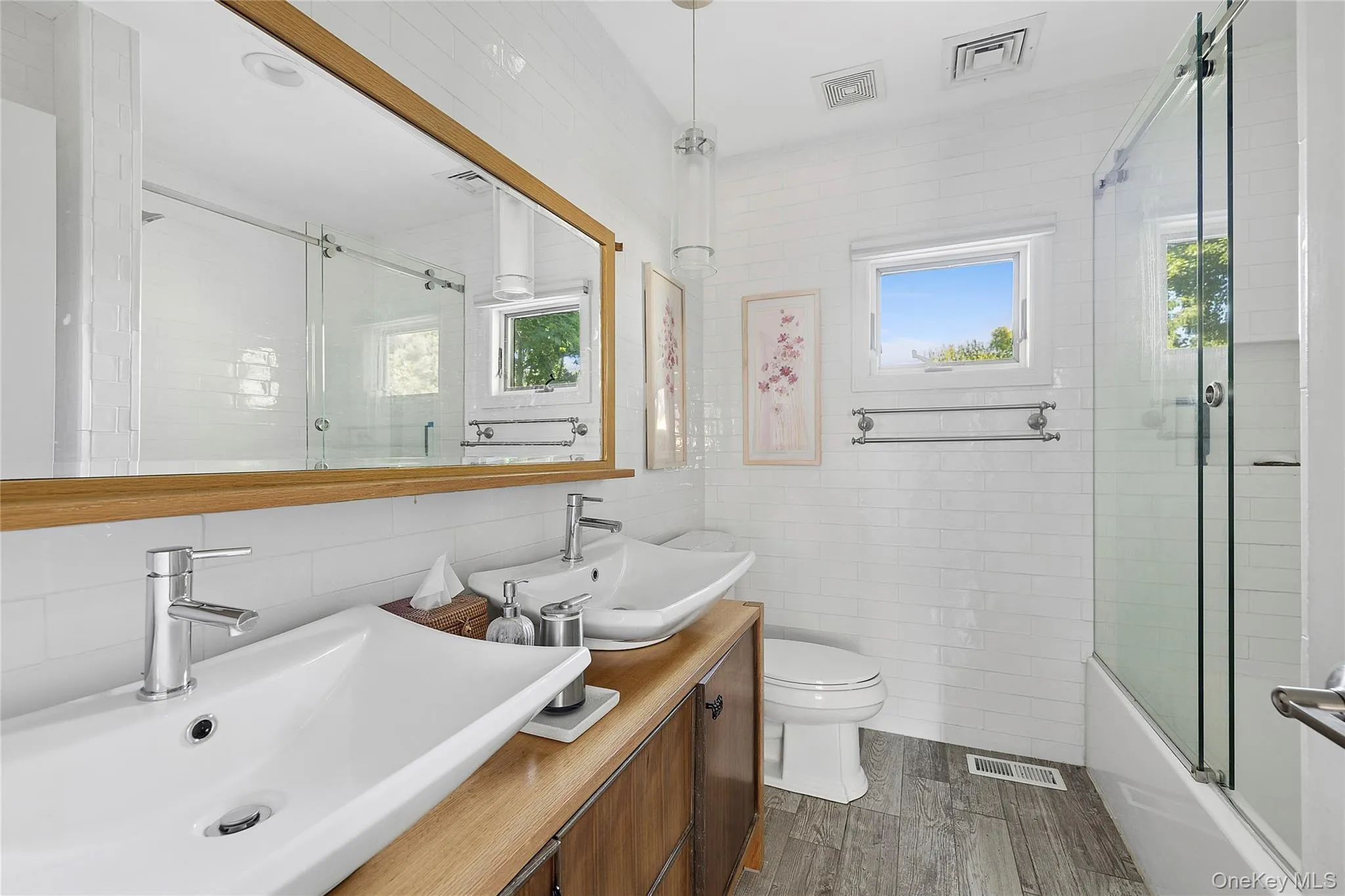 Bathroom featuring a dual vessel sink vanity with wood-finish countertop, wood-finish flooring, white subway tile walls, and a glass-enclosed shower-tub combination Bathroom featuring a dual vessel sink vanity with wood-finish countertop, wood-finish flooring, white subway tile walls, and a glass-enclosed shower-tub combination
