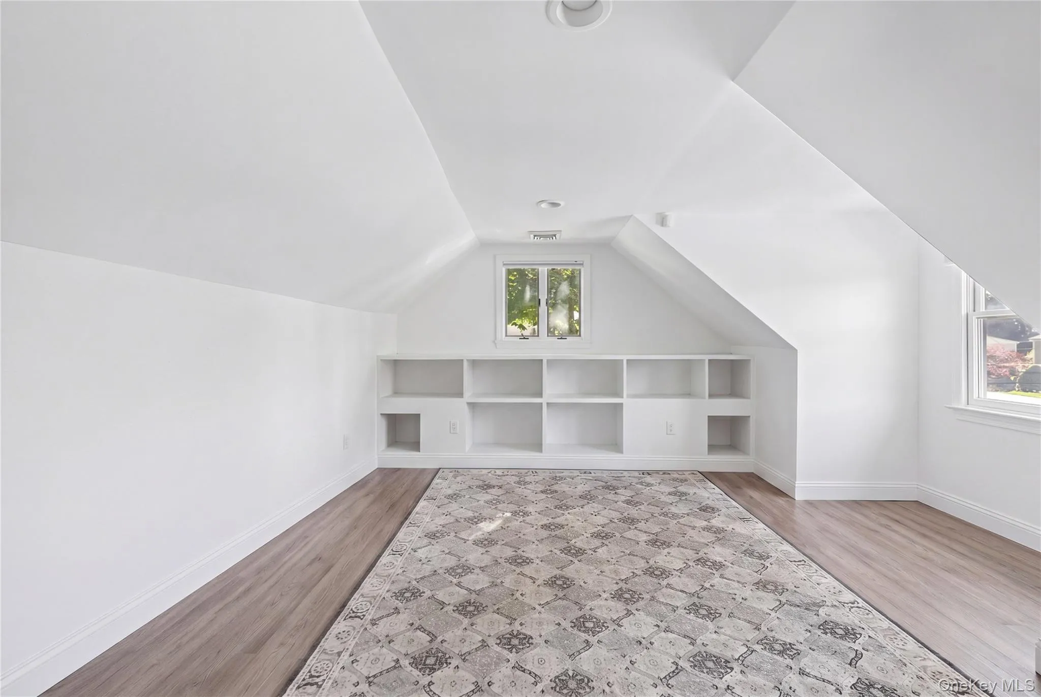 Bright room with wood-finish flooring, built-in shelving, two windows, recessed lighting, and sloped ceilings Bright room with wood-finish flooring, built-in shelving, two windows, recessed lighting, and sloped ceilings